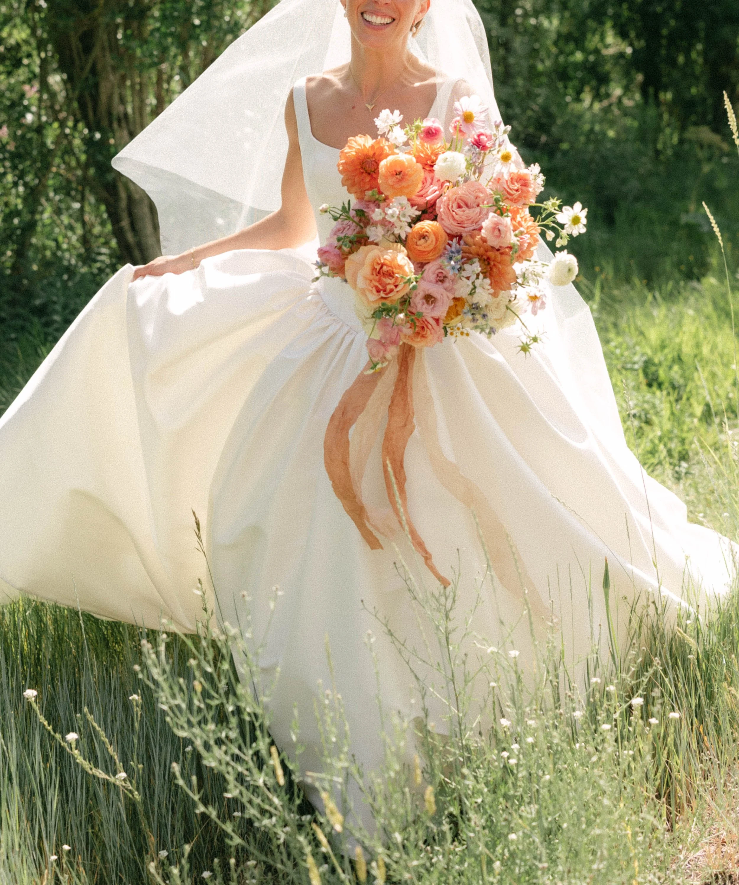 A bride in a white wedding dress holding a large bouquet of colorful flowers, standing outdoors in a grassy area with trees in the background.