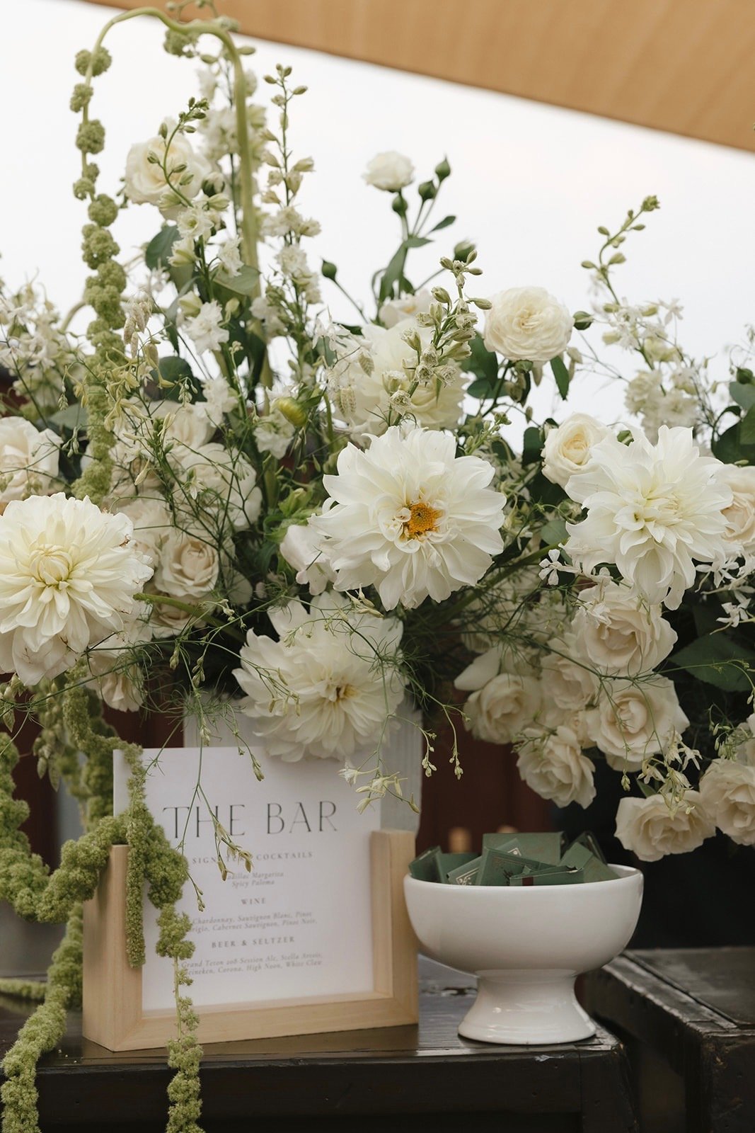 A floral arrangement of white roses and dahlias on a wooden table, with a sign that says 'The Bar' and a bowl filled with small green packages.