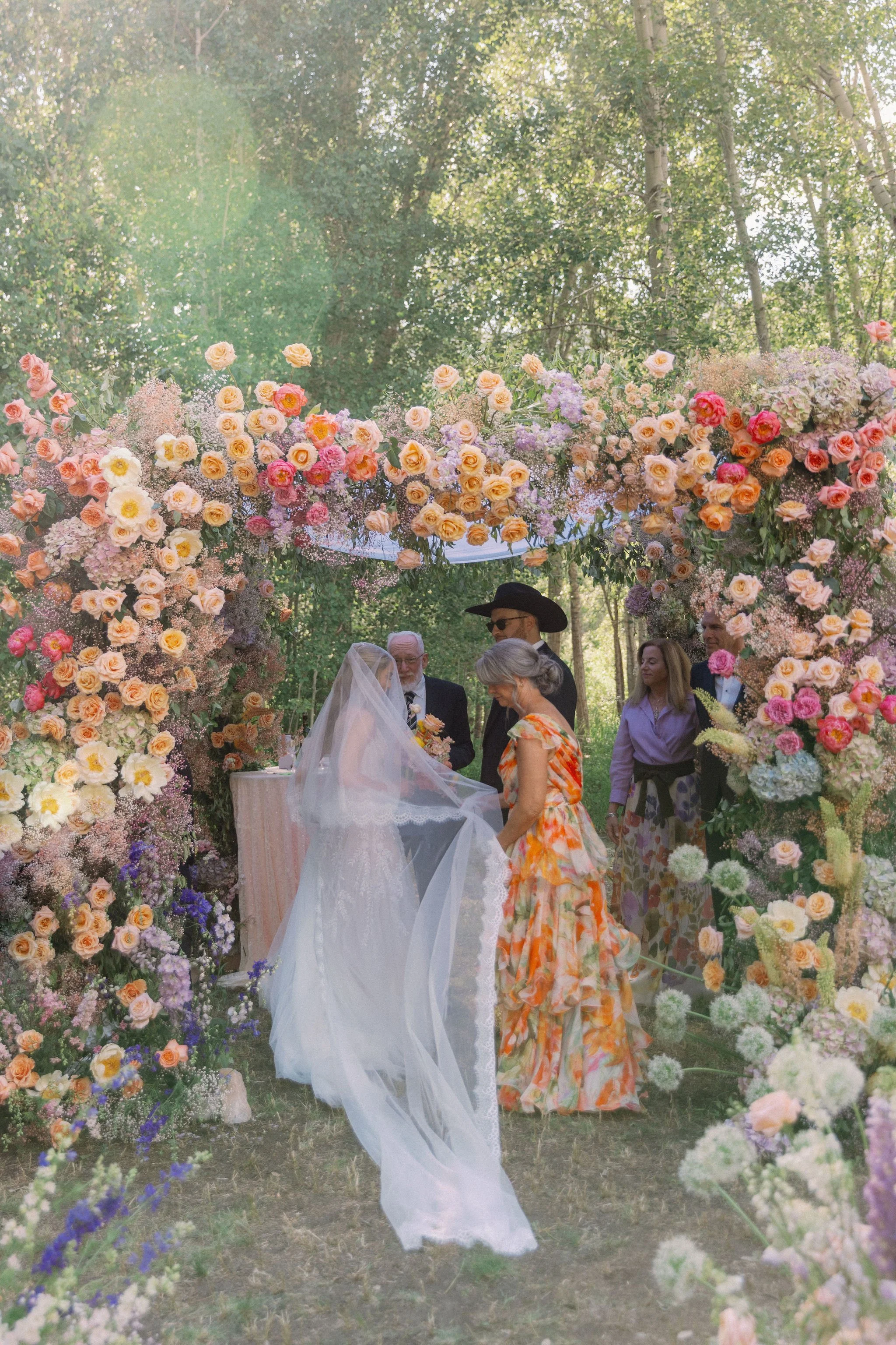 A wedding ceremony outdoors under a floral arch with pink, peach, and purple flowers, with a bride and groom facing each other, surrounded by guests in a wooded area.