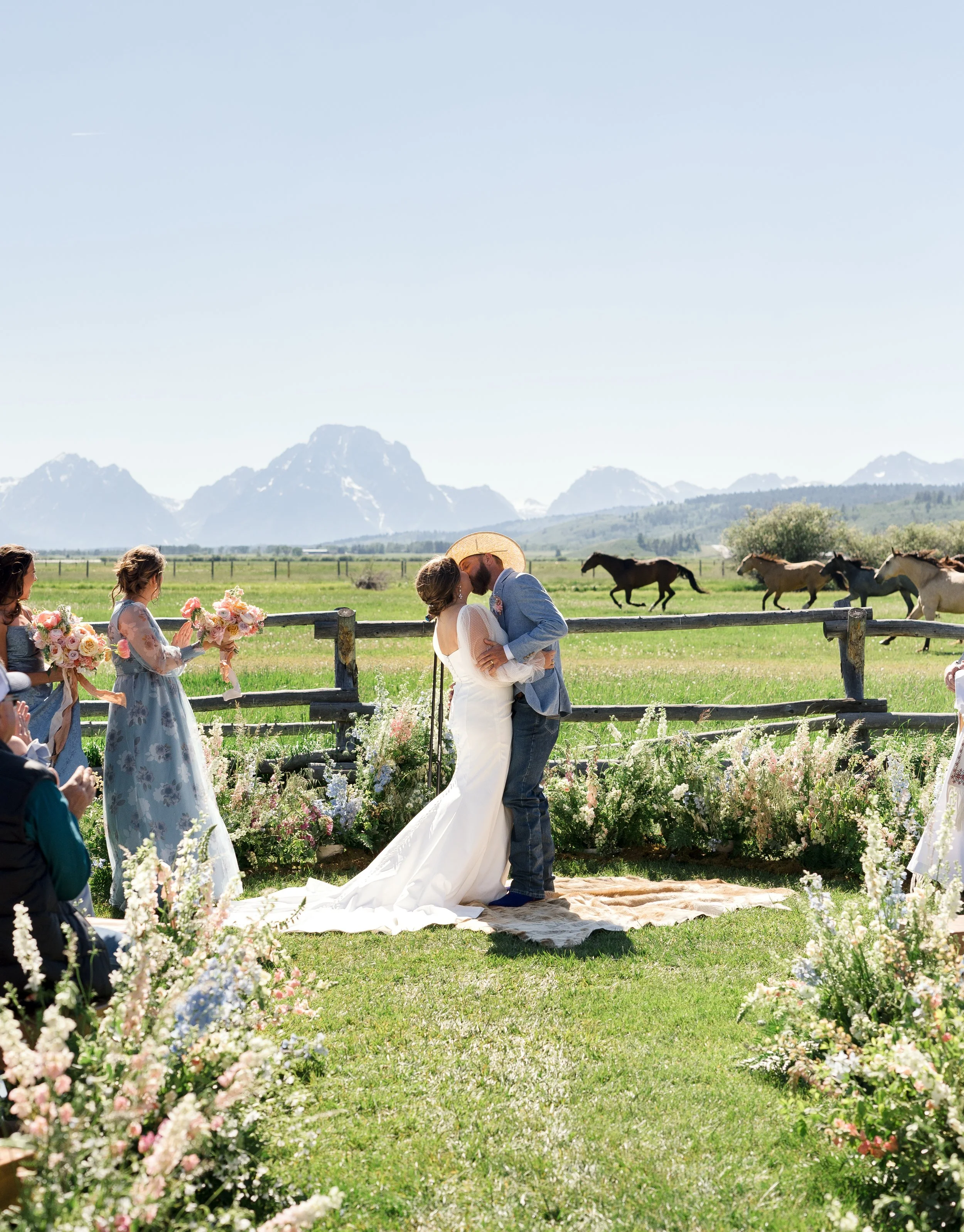A couple getting married outdoors with mountains in the background and horses in the field. They are kissing, with bridesmaids holding bouquets on the side and guests seated. The scene is decorated with flowers.