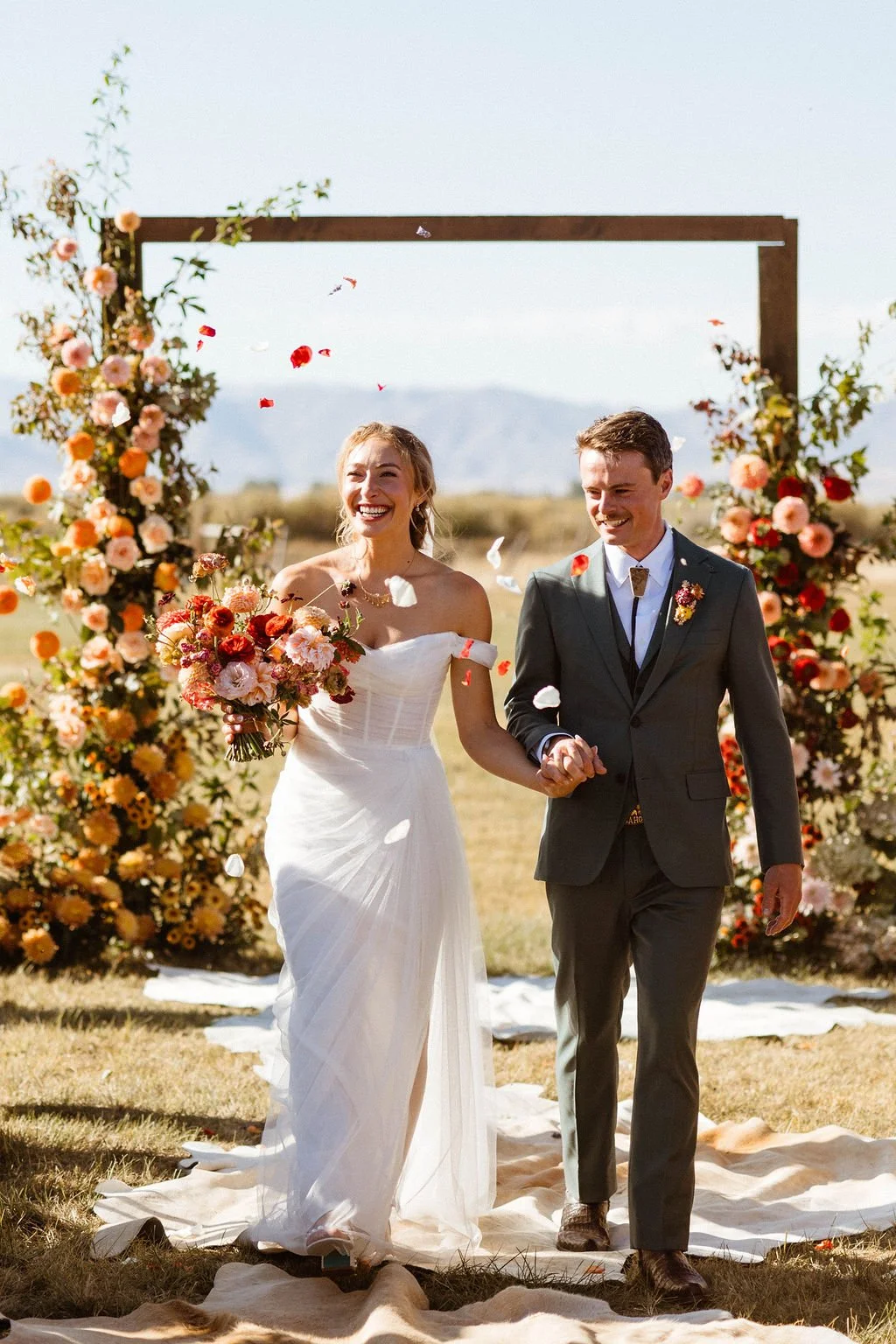 A newlywed couple walking hand in hand under a floral wedding arch in an outdoor setting. The bride is holding a bouquet and wearing a white wedding dress, while the groom is in a dark suit. Flower petals are falling around them, and they are smiling