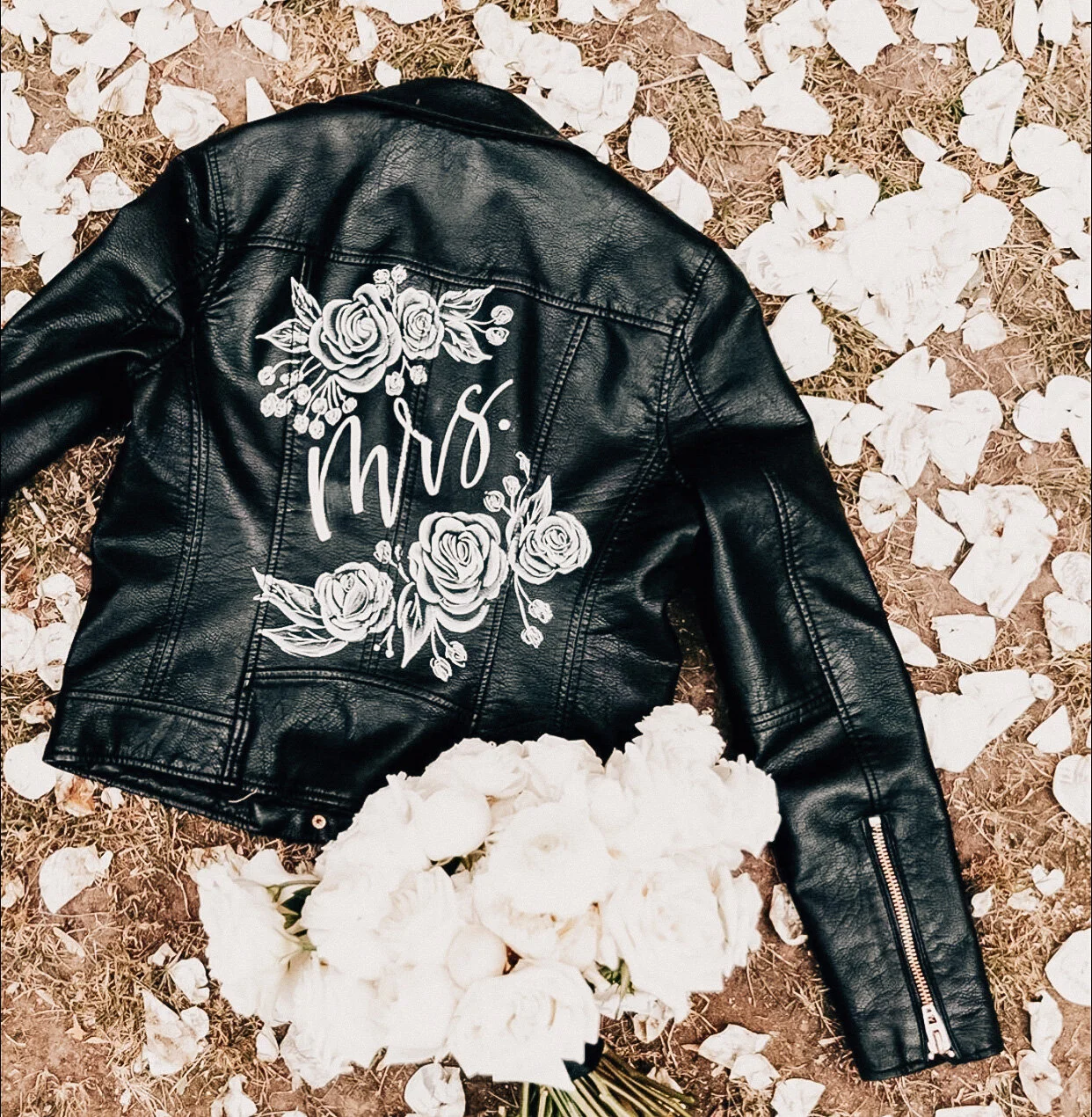 A black leather jacket with floral embroidery and the word 'Mrs.' on the back, lying on the ground amid white flower petals.