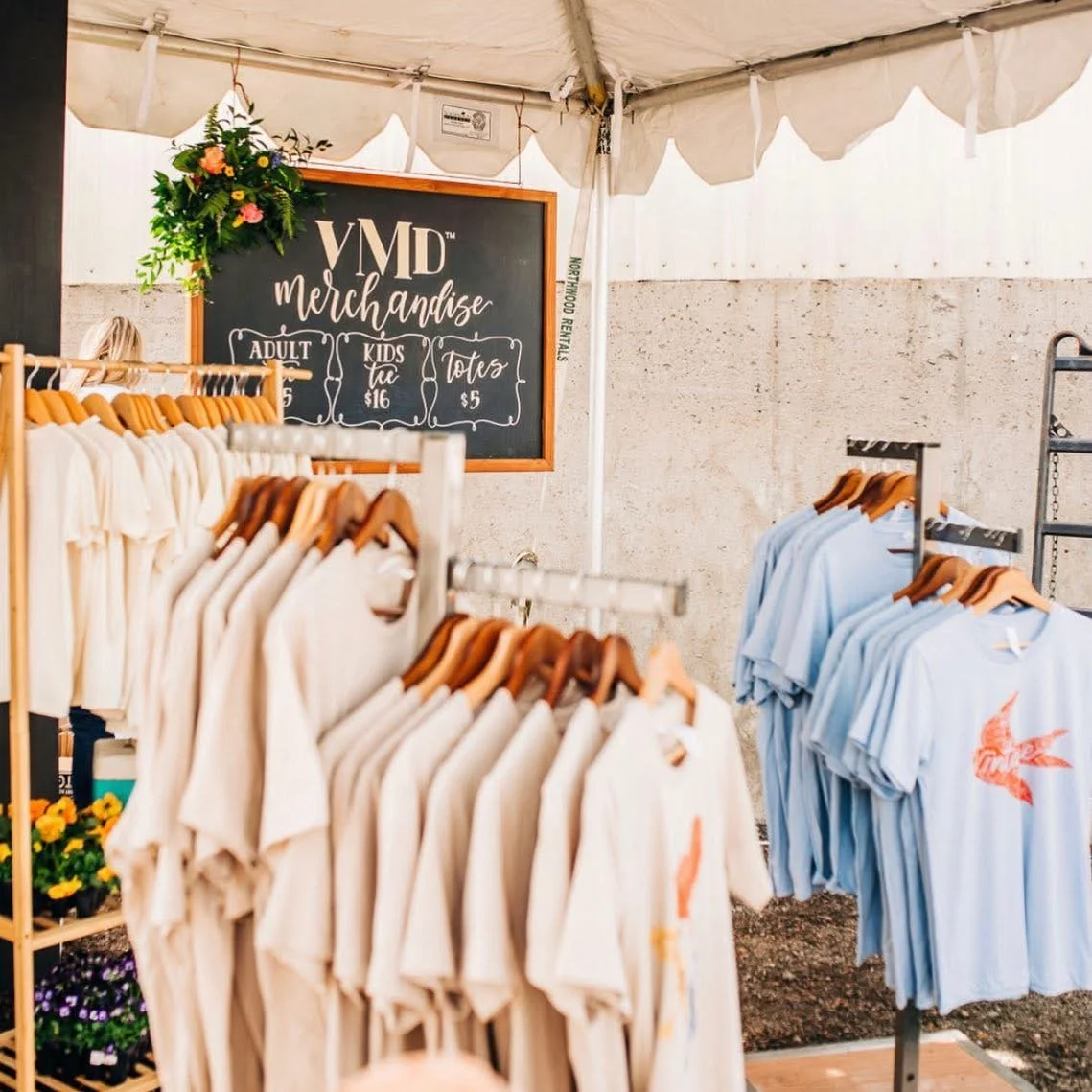 A merchandise booth with t-shirts hanging on racks and a blackboard sign displaying prices for adult, kids' ice, and totes, with flowers and a tent overhead.