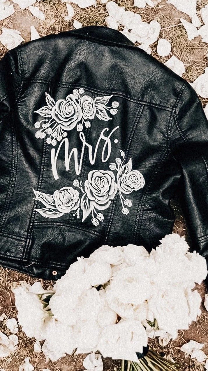 Black leather jacket with white floral designs and the word 'Mrs' written on the back, lying on the ground among white flower petals, with a bouquet of white flowers in the foreground.