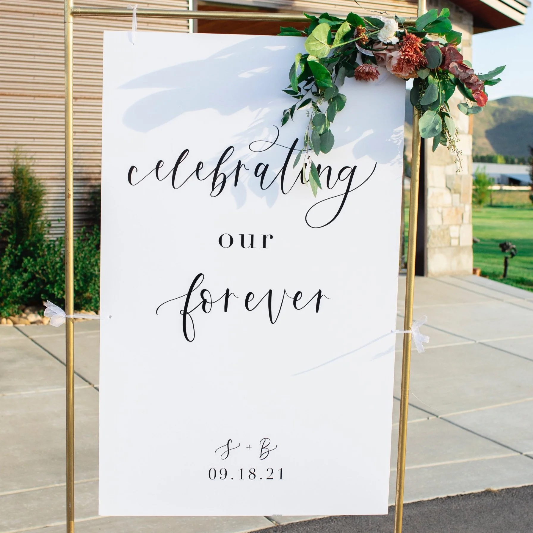 Wedding sign with floral decoration, reading 'celebrating our forever' with initials 'S + B' and date '09.18.21'.