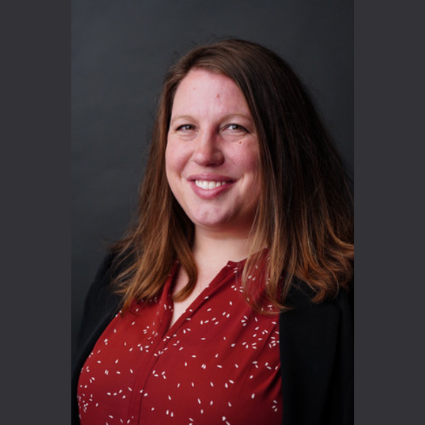 [Image description (provided by the person pictured):  Photo of Kari Slater, a smiling young fat white female with shoulder-length light brown hair, wearing a black sweater over a patterned dress.]