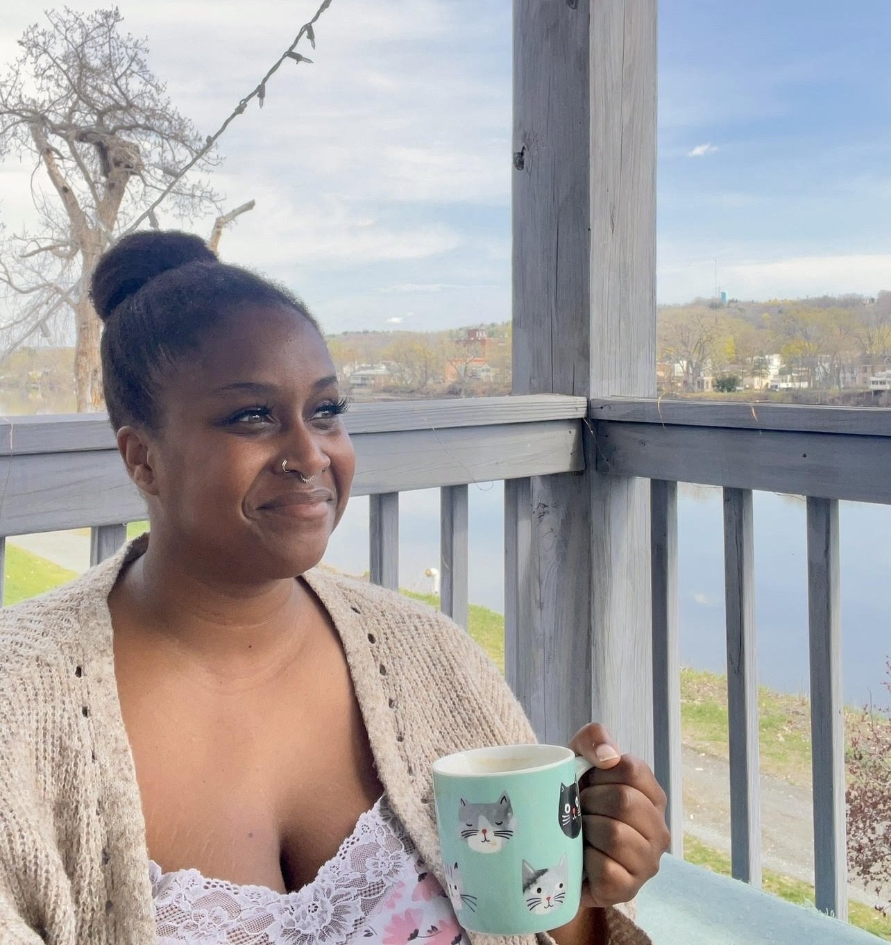 A headshot of Kara Rabii, a smiling Black woman with medium-brown skin and dark hair in a high bun, wearing a cream cardigan over a light lace top and holding a light blue bug on a balcony overlooking the water.