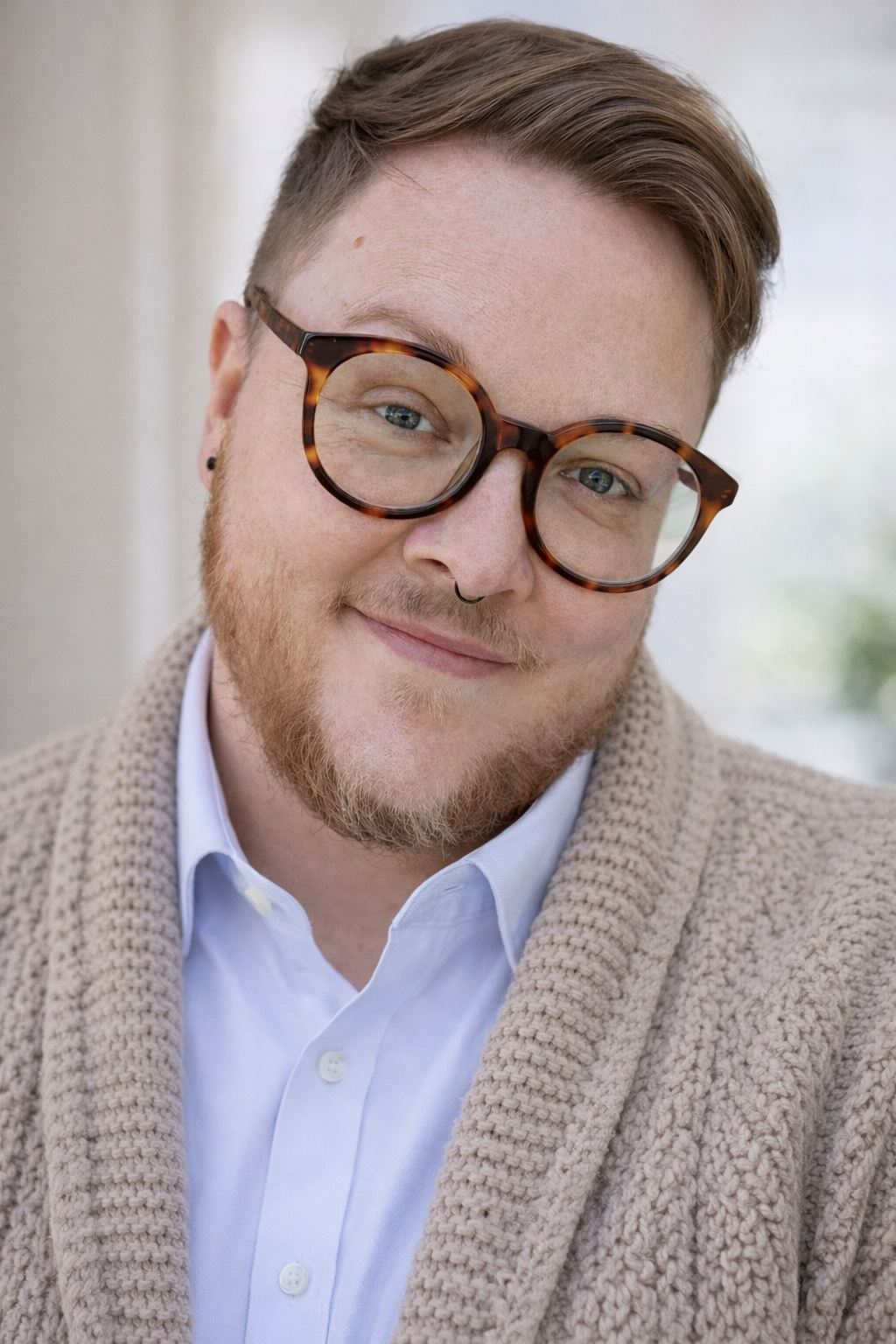headhsot of Vaughn Darst, a white transmasculine man with blonde hair, beard, framed round glasses, and a black septum ring. He is wearing a blue collared shirt and a tan sweater.