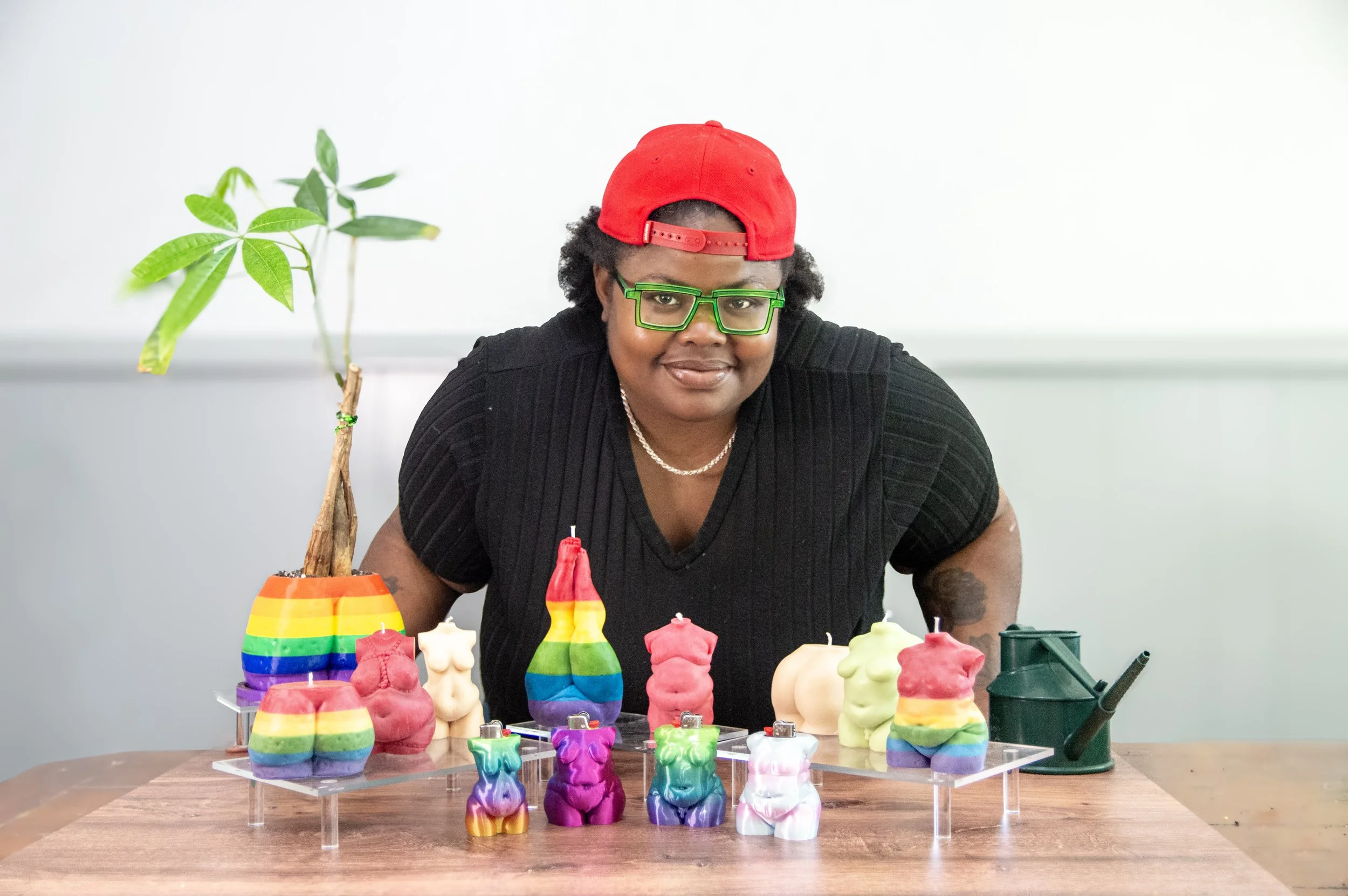 Photo of Jodyann Morgan standing behind a table wearing green glasses, a red hat, and a black shirt. She's leaning forward over body shaped candles, planters, and lighter holders from CTOAN, her fat art company.