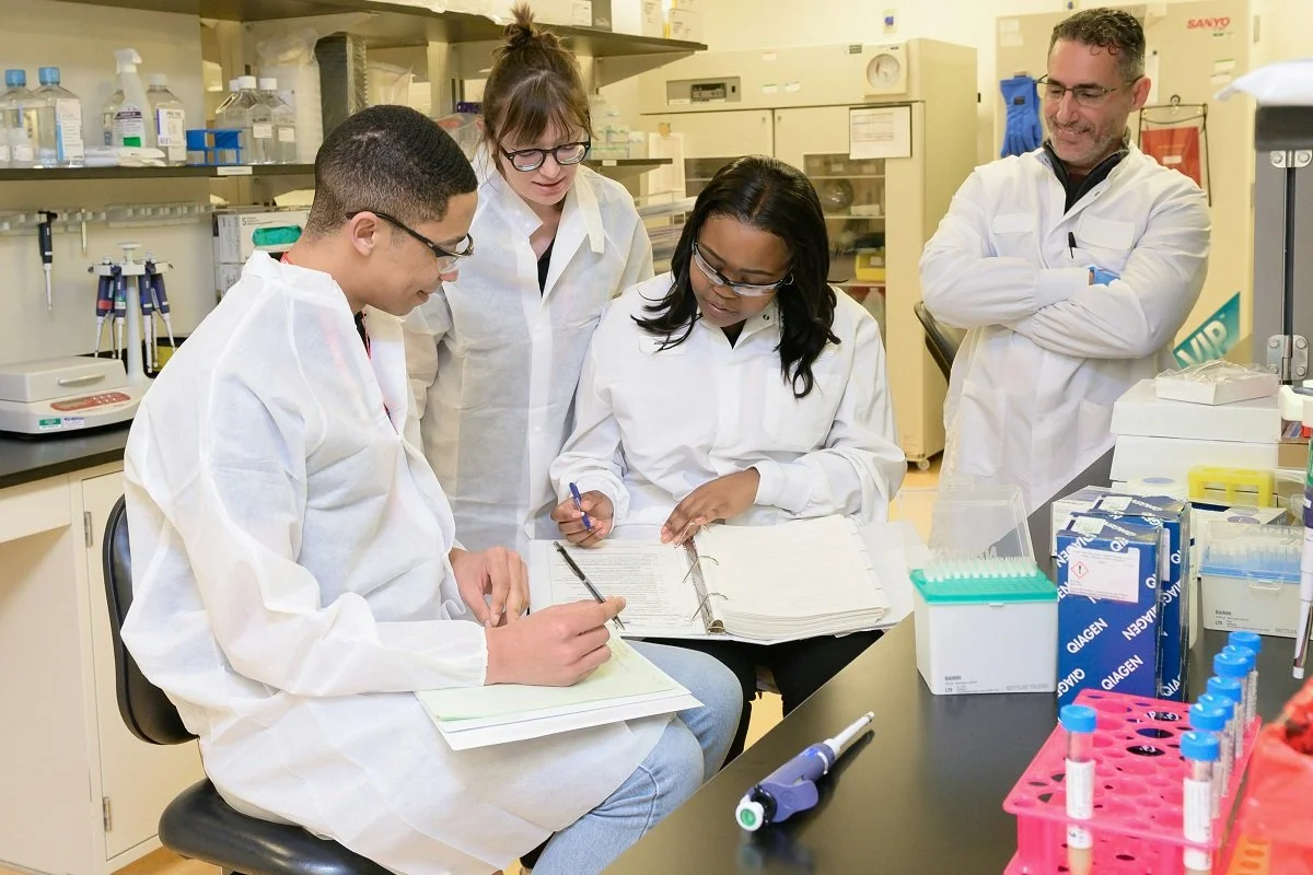 [Accessibility description: A photo of four people in a laboratory, all wearing white coats, gathered around notebooks, appearing in mid-discussion.]