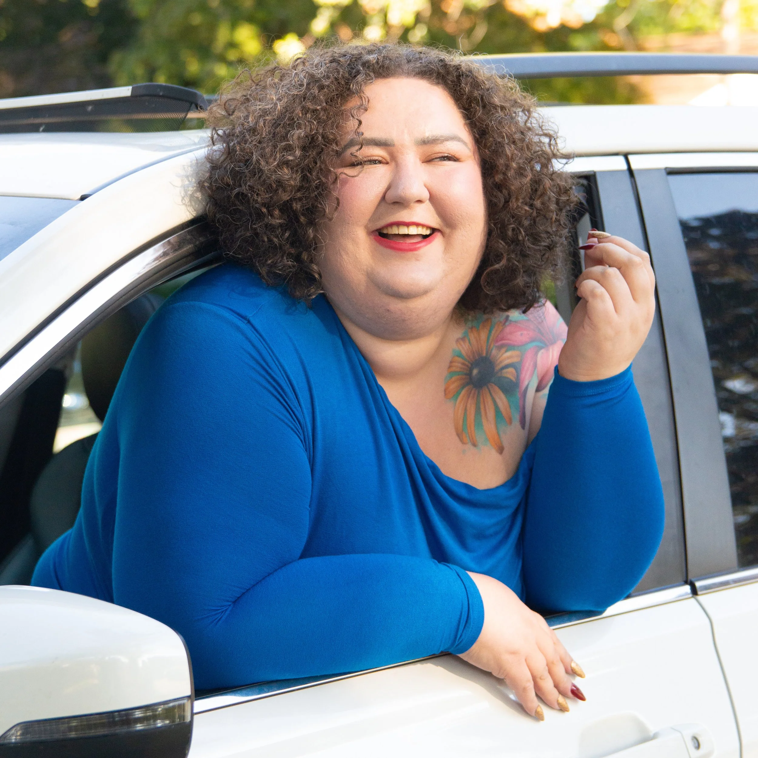 A photo of Chaya M Milchtein, a fat white woman with curly hair in a blue dress photographed through the driver's window of her car that she's leaning out of.