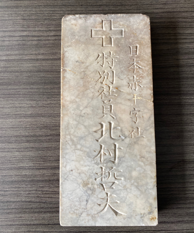 A rectangular stone with a carved cross at the top and Japanese characters engraved below, placed on a wood surface. It is referred to as the Nagasaki Marble.