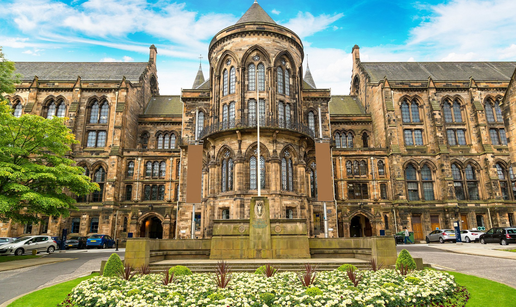 Front view of a University of Glasgow Medical School, a historic stone Gothic-style building with arched windows, a central tower, and a fountain with flowers in front.