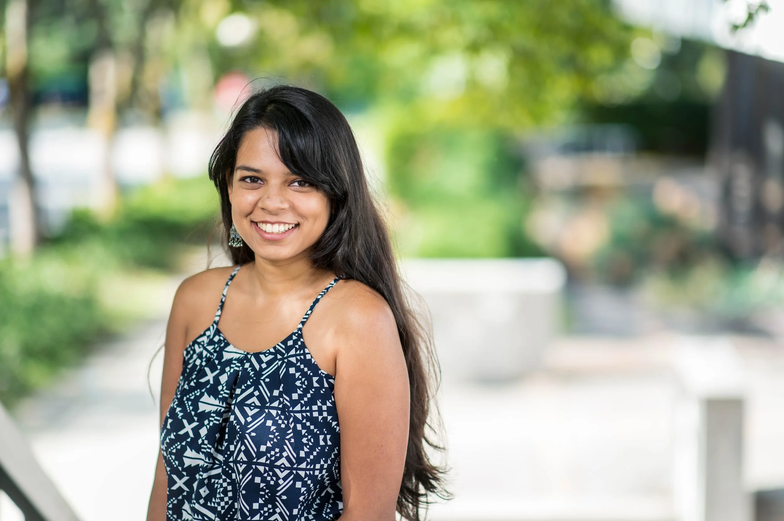 A woman with long dark hair smiling outdoors with green trees and a pathway in the background.