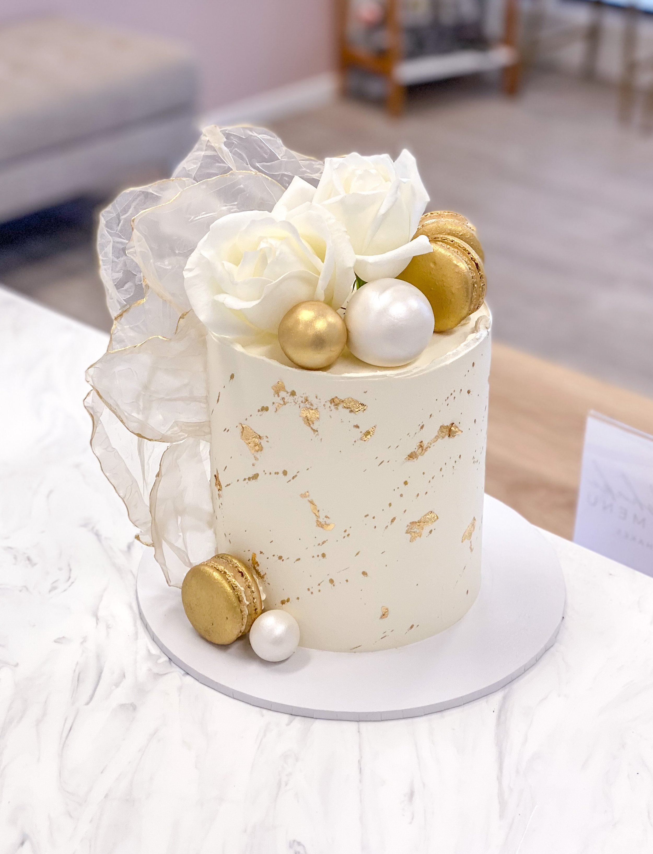 Elegant white cake with gold accents, topped with white roses, gold and white spherical decorations, and gold macarons, on a marble surface.