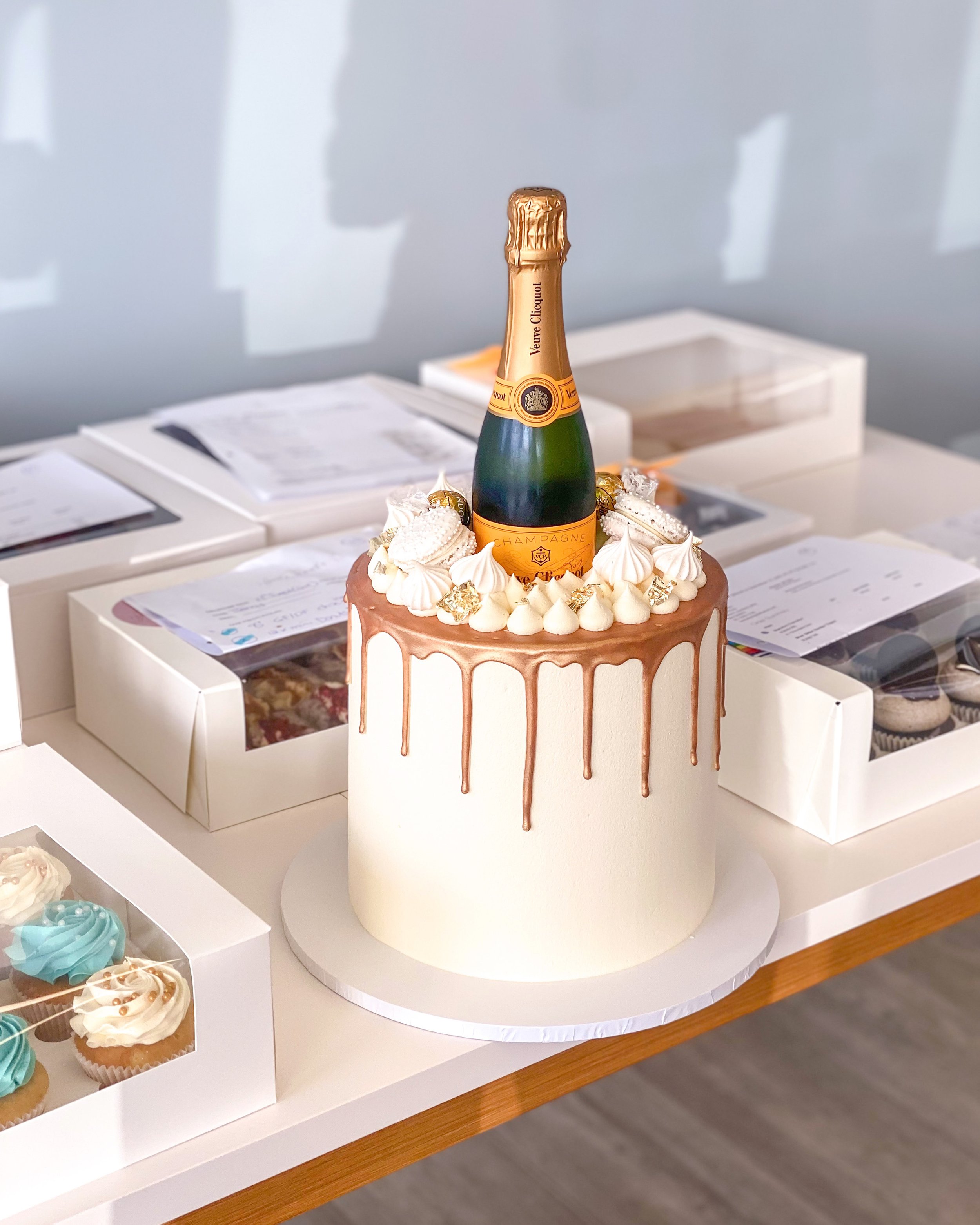 Tall celebration cake with chocolate drip and a bottle of champagne on top, surrounded by boxed baked goods on a table.