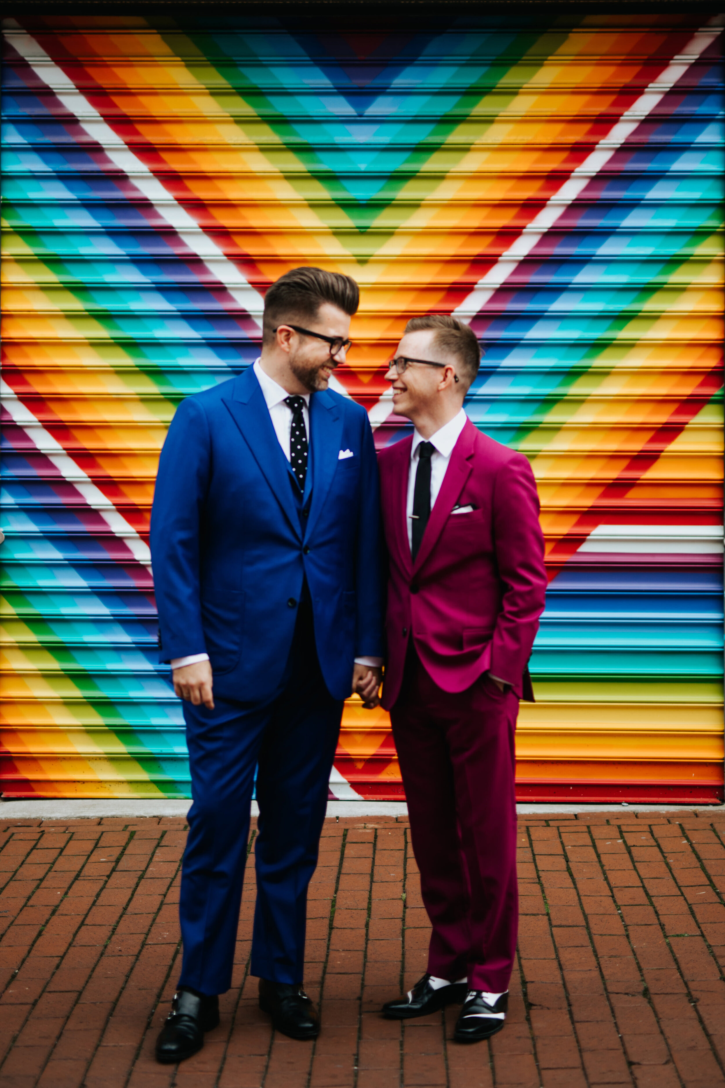 LGBTQ couple in blue and maroon suits posing by a rainbow mural in NYC Shawnee Custalow Queer Wedding Photographer