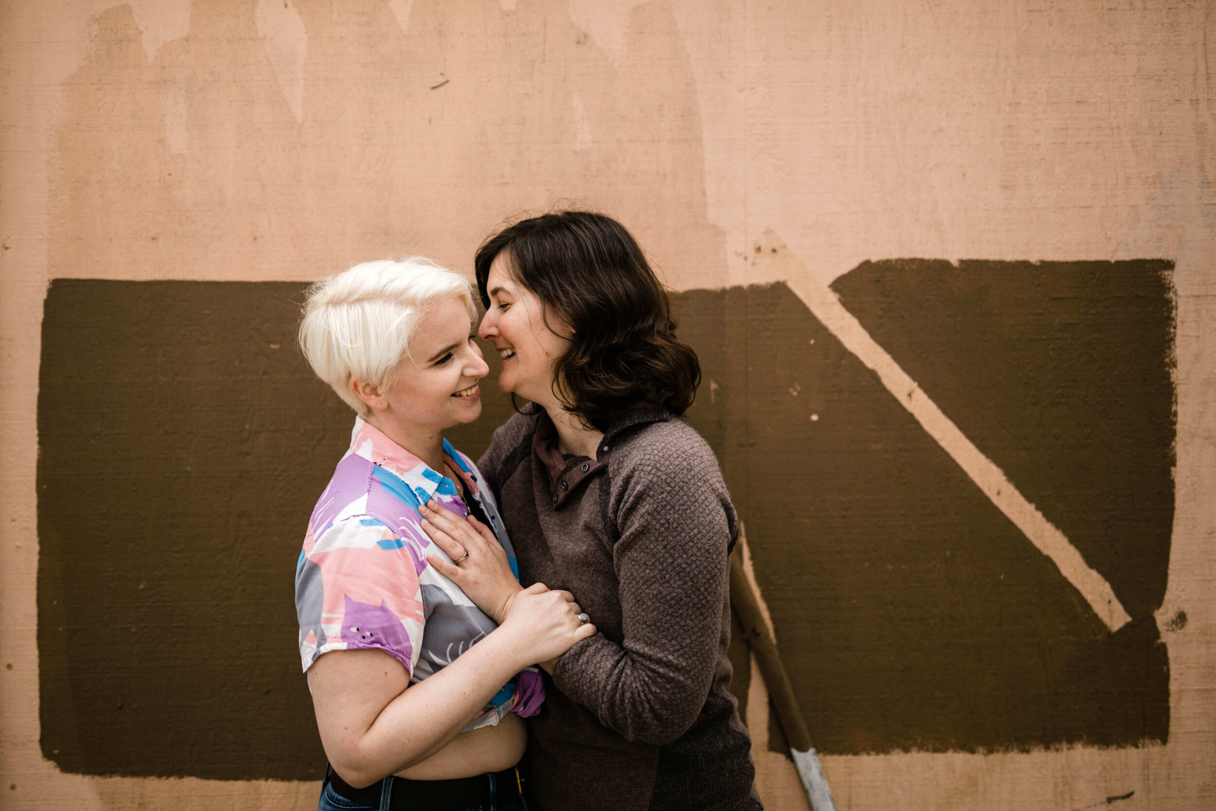 Engaged queer couple smiling and embracing by a brown painted rectangle Shawnee Custalow DC Wedding Photographer