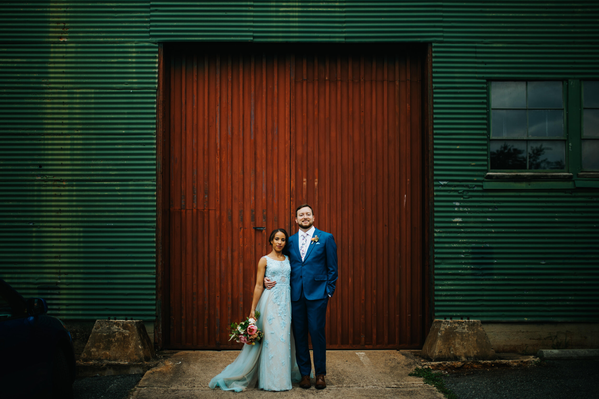 Bride and groom wearing blue posing by a red and green warehouse RVA Shawnee Custalow Queer Wedding Photographer