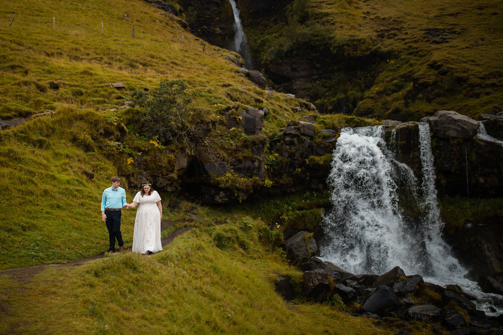 Newlywed couple walking along a path by a waterfall in Iceland Shawnee Custalow Queer Wedding Photographer