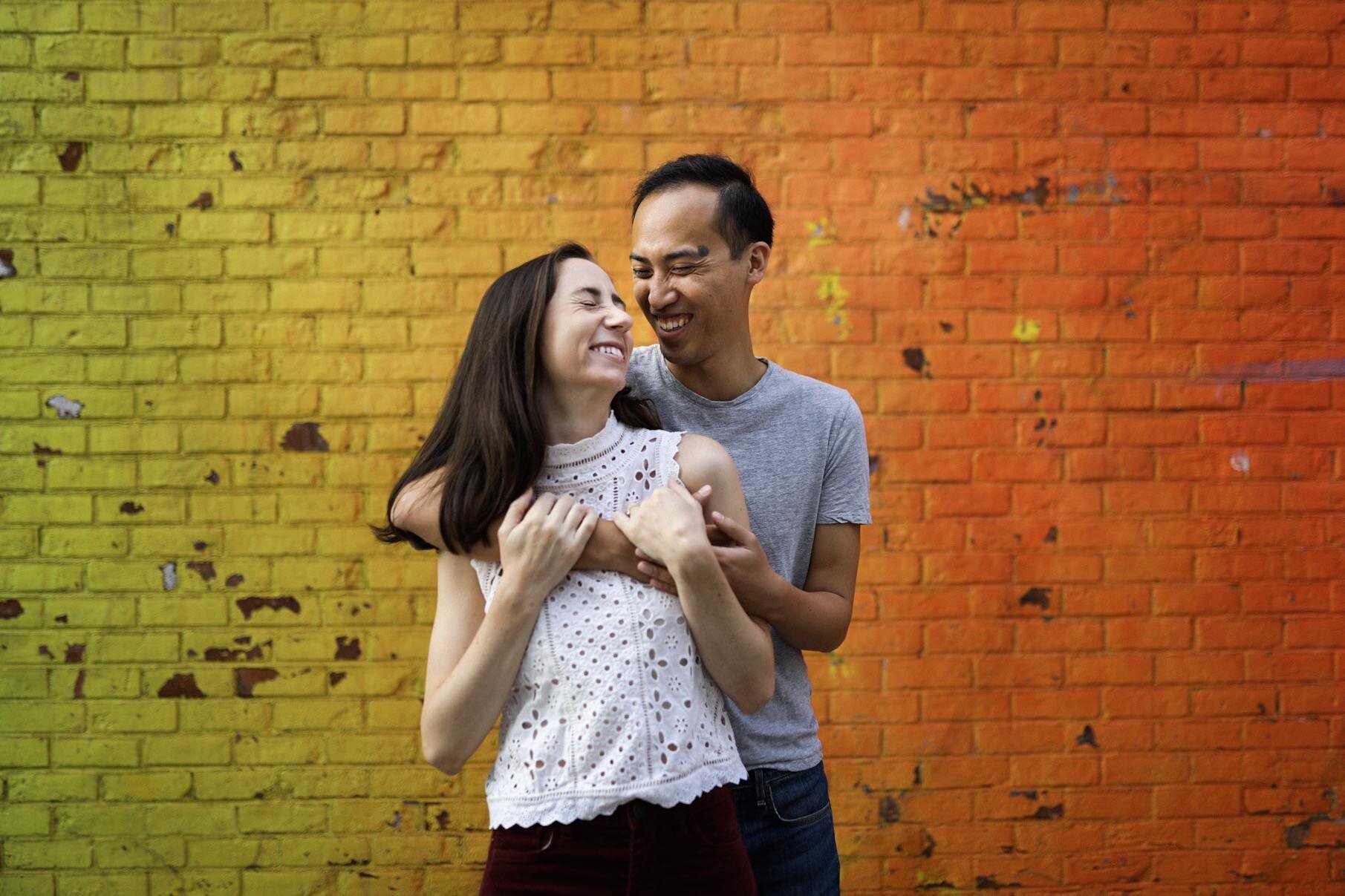 Engaged couple embracing and laughing by a colorful mural in Richmond VA Shawnee Custalow Queer Wedding Photographer