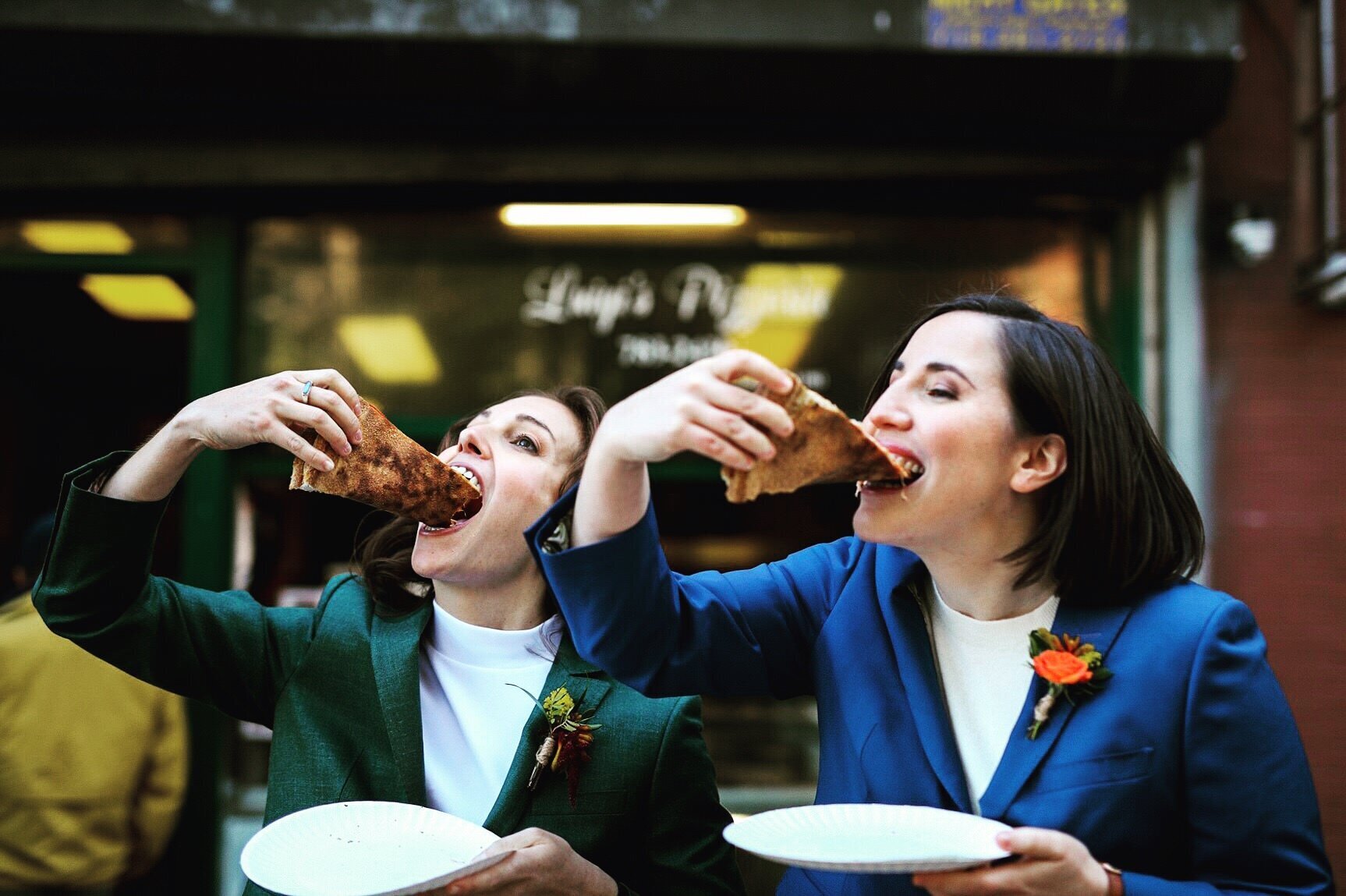 Two brides in matching suits eating slices of pizza in Richmond VA Shawnee Custalow Queer Wedding Photographer