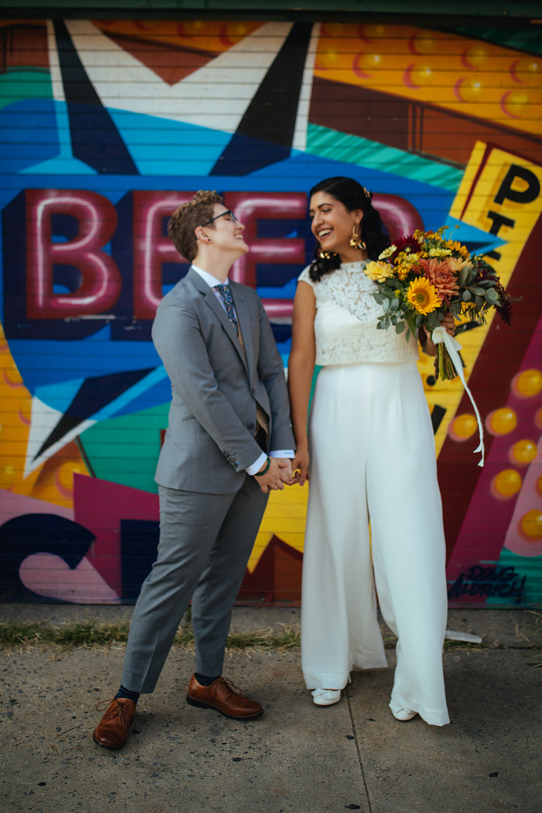 LGBTQ holding hands and smiling by a mural in Red Hook Brooklyn Shawnee Custalow Queer Wedding Photographer