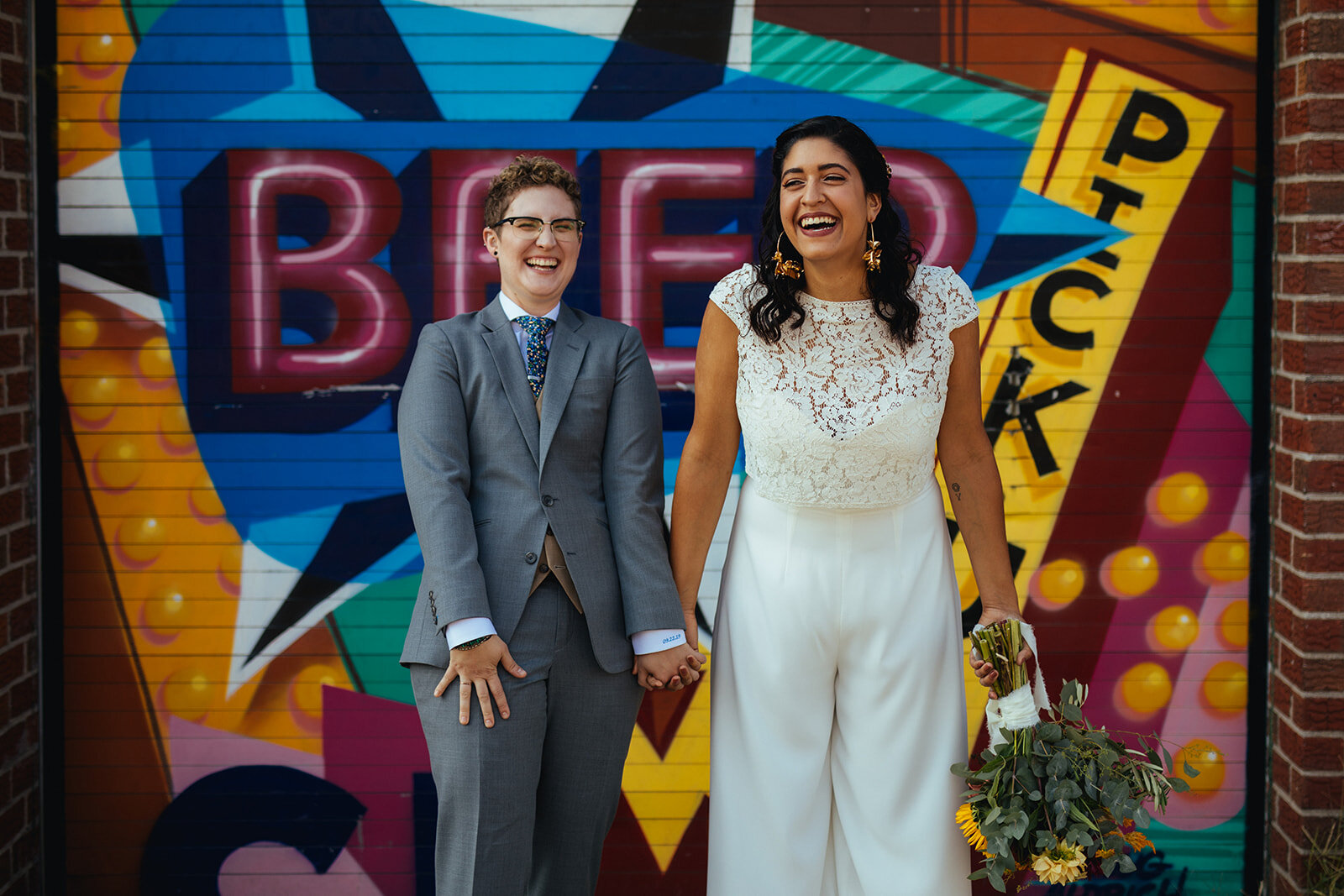 LGBTQ couple laughing and holding hands before a mural in Red Hook Brooklyn Shawnee Custalow Queer Wedding Photographer