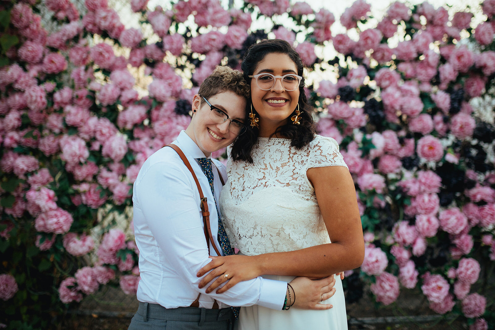 LGBTQ newlywed couple embracing by big pink flowers in Brooklyn NY Shawnee Custalow Queer Wedding Photographer