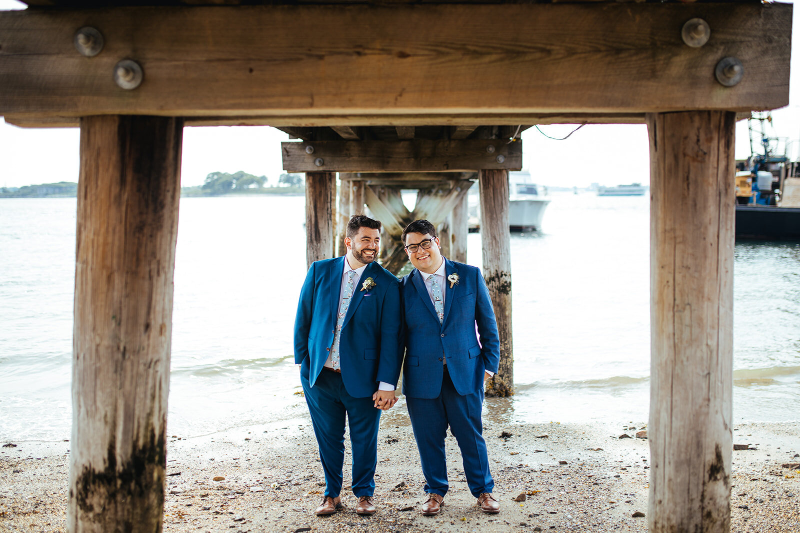 Grooms holding hands under a pier in Portland ME Shawnee Custalow Queer wedding photography