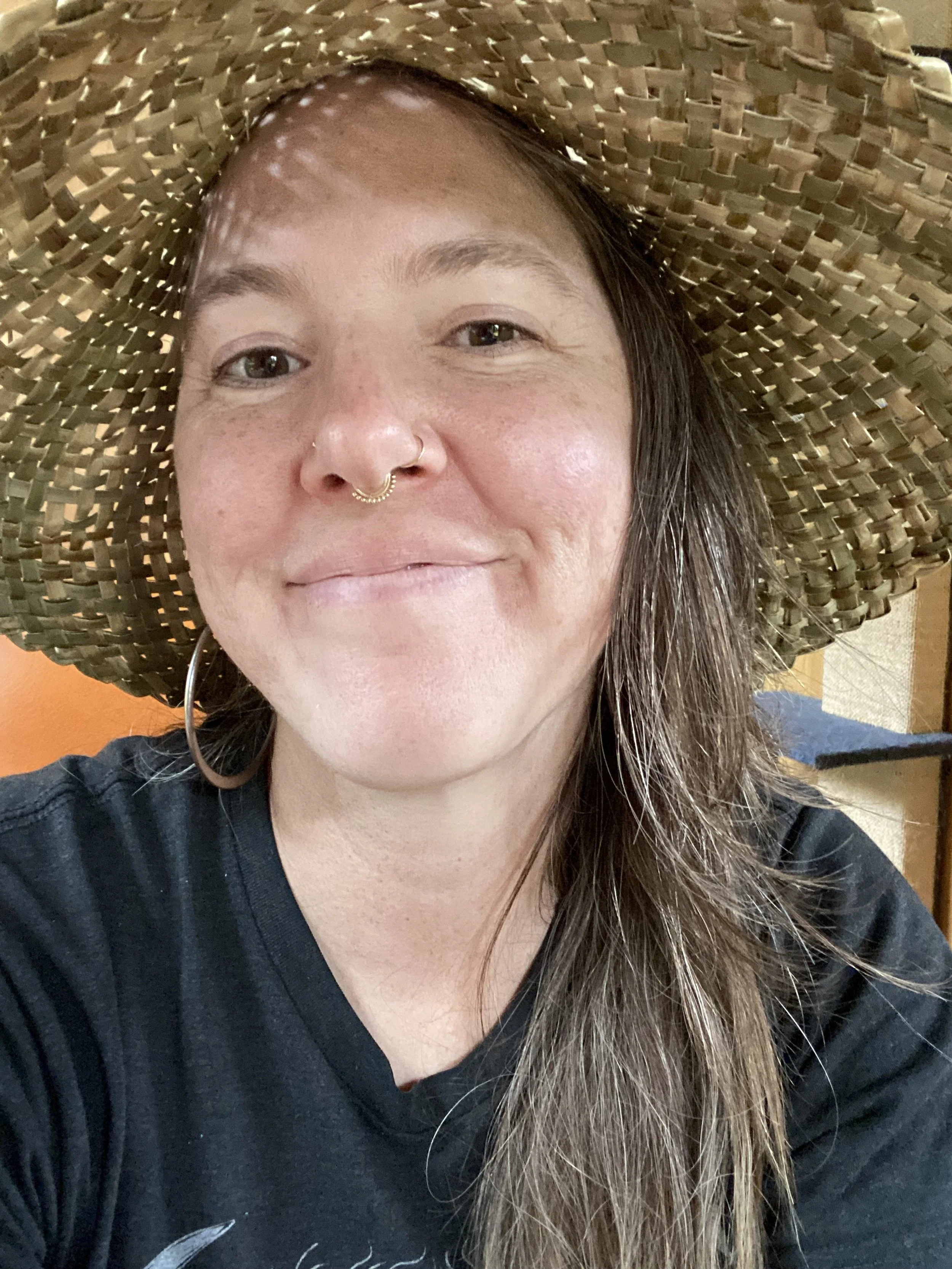 A woman taking a selfie, wearing a large woven sun hat, with long brown hair, earrings, and a septum piercing, smiling in a casual setting.