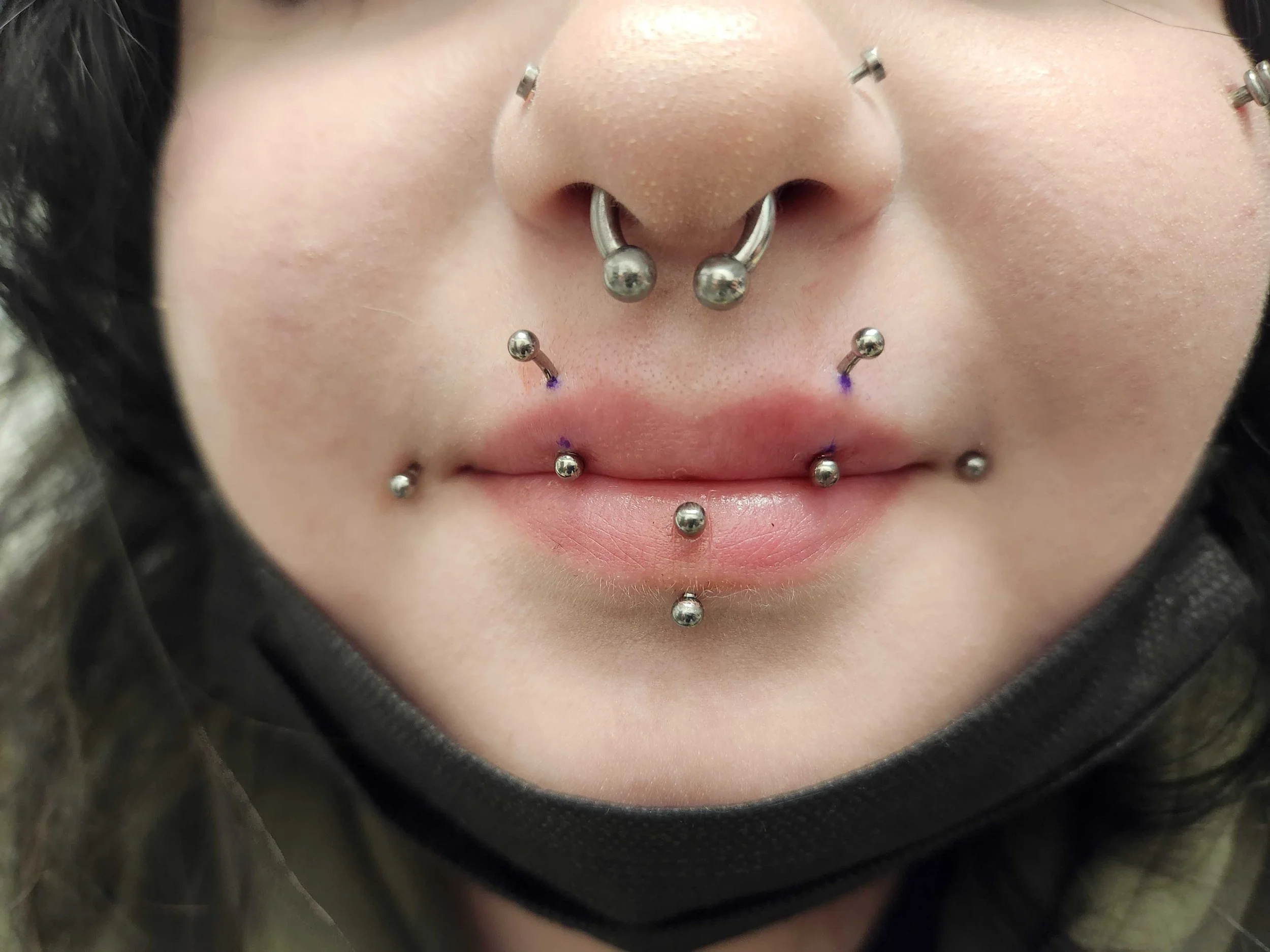 Close-up of a person's face with multiple facial piercings, including lip, septum, and cheek piercings.