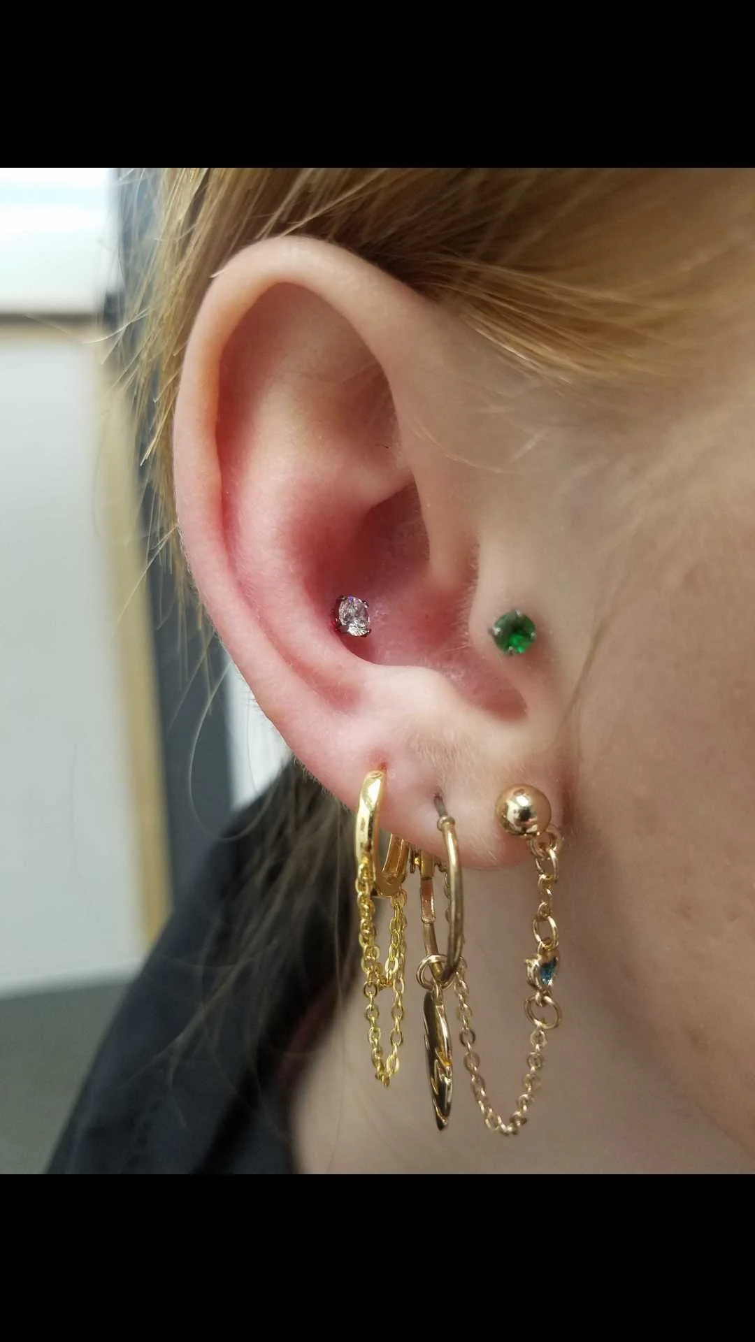 Close-up of an ear with multiple piercings, including a diamond stud, green gemstone, and gold hoop earrings with chains.