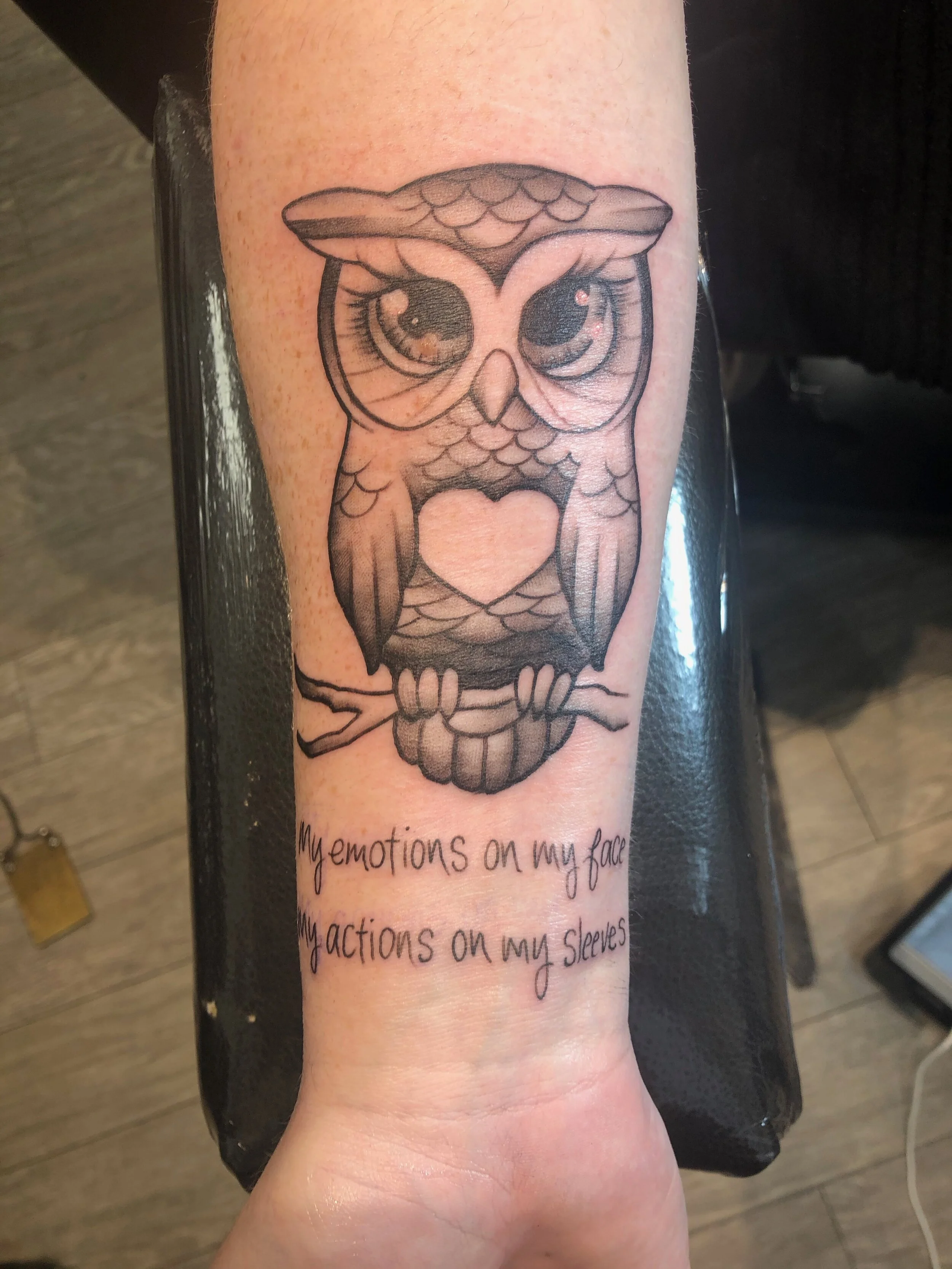 Tattoo of an owl with a heart on its chest, perched on a branch. Below, there is text saying "my emotions on my face, my actions on my sleeves."