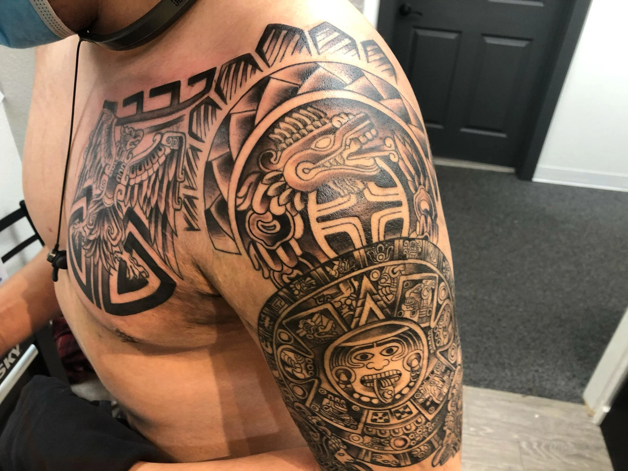 Person with detailed Aztec-inspired tattoos on shoulder and chest, featuring symbols and patterns.