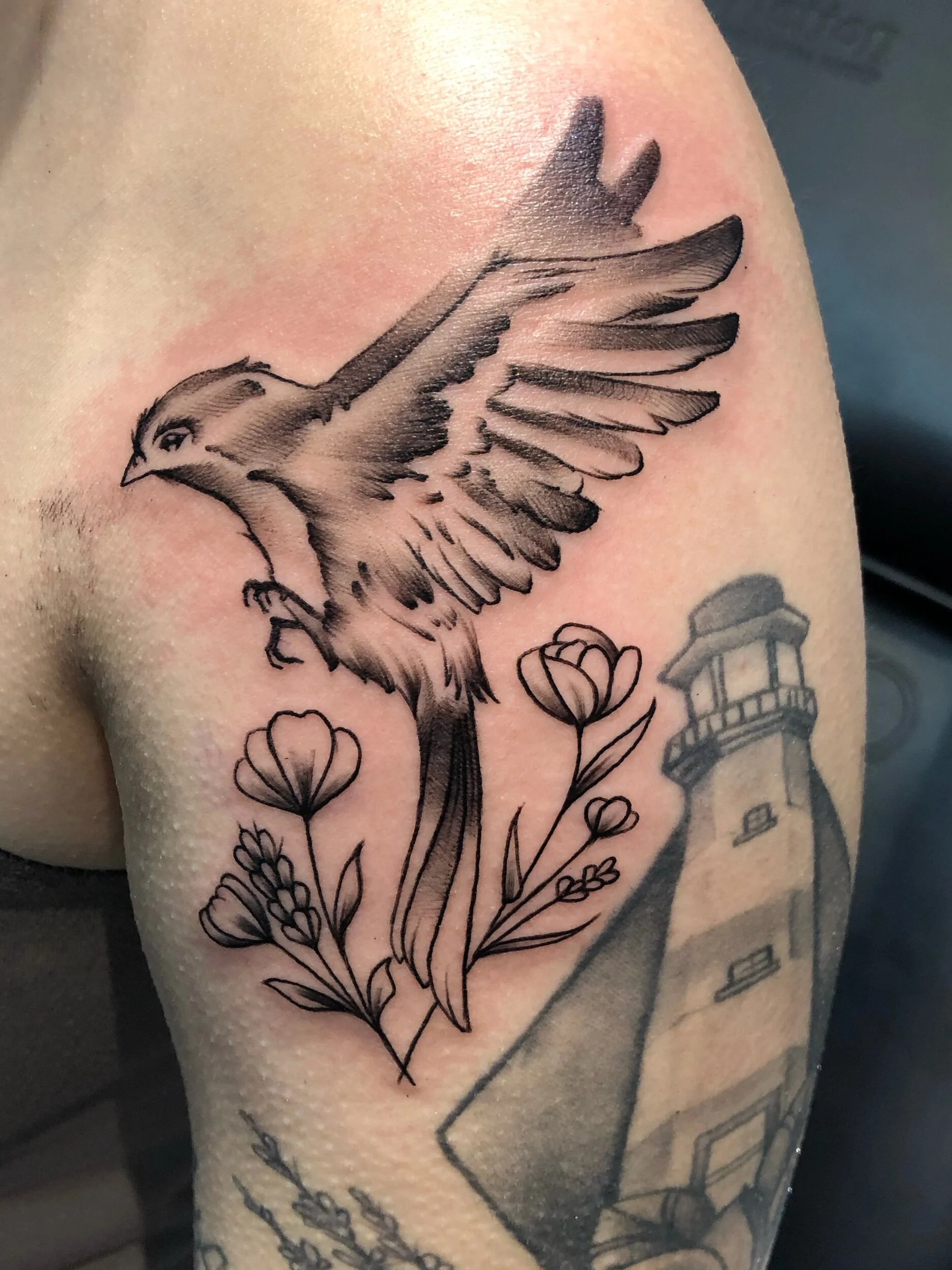 Tattoo of a bird in flight with flowers on an arm, featuring a lighthouse in the background.