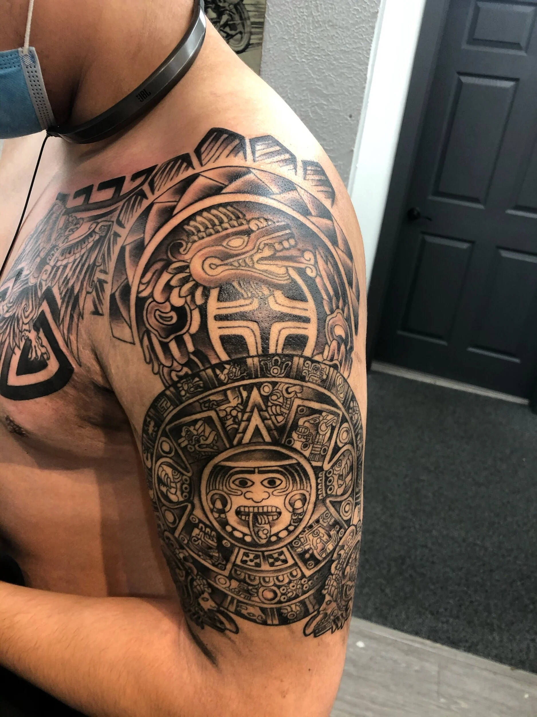 A detailed black tattoo on a person's upper arm featuring Aztec or Mayan artwork, including a sun or calendar symbol surrounded by intricate patterns.