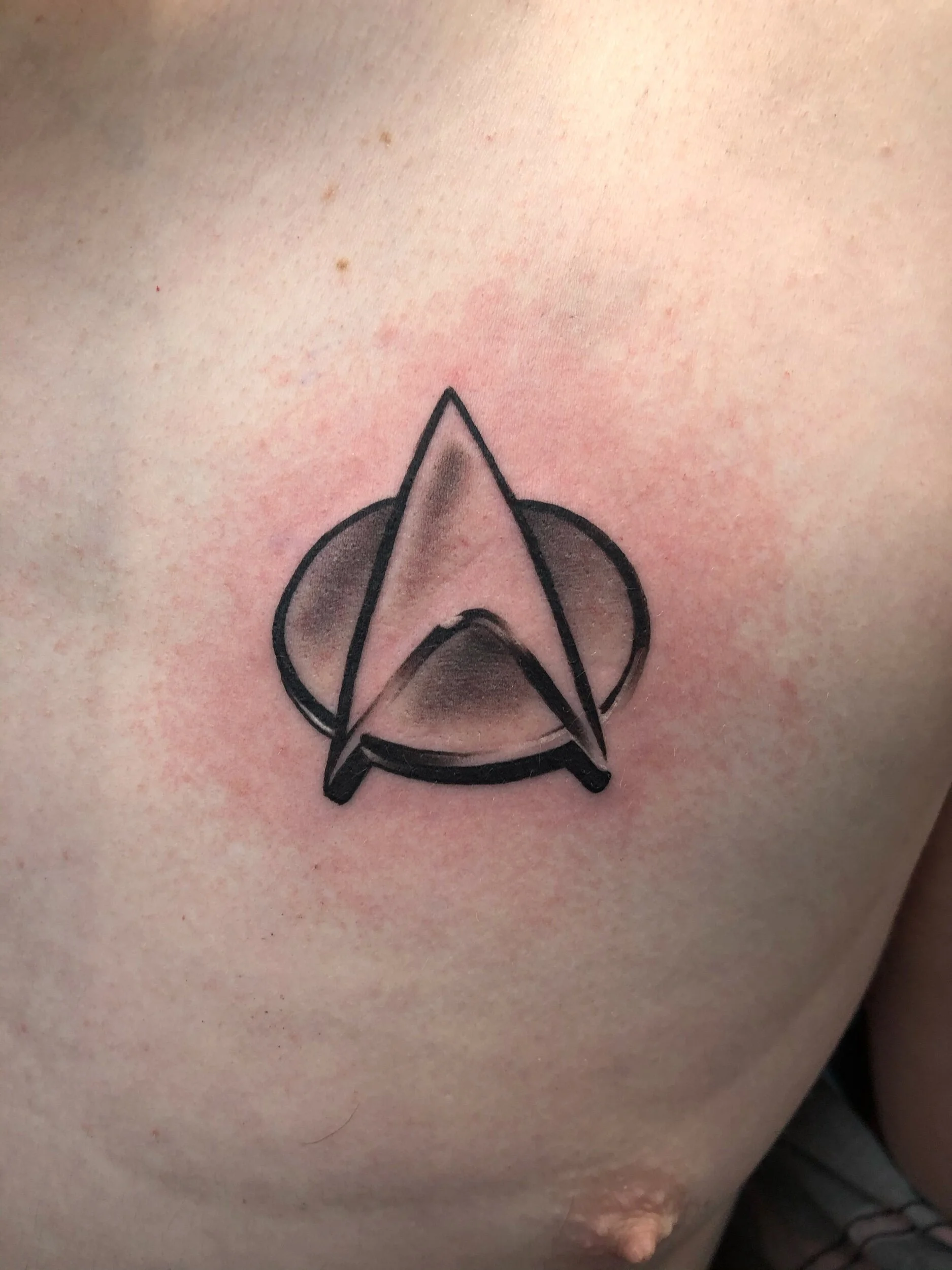 Tattoo of a stylized insignia on a person's chest featuring a triangular shape over a circular background.