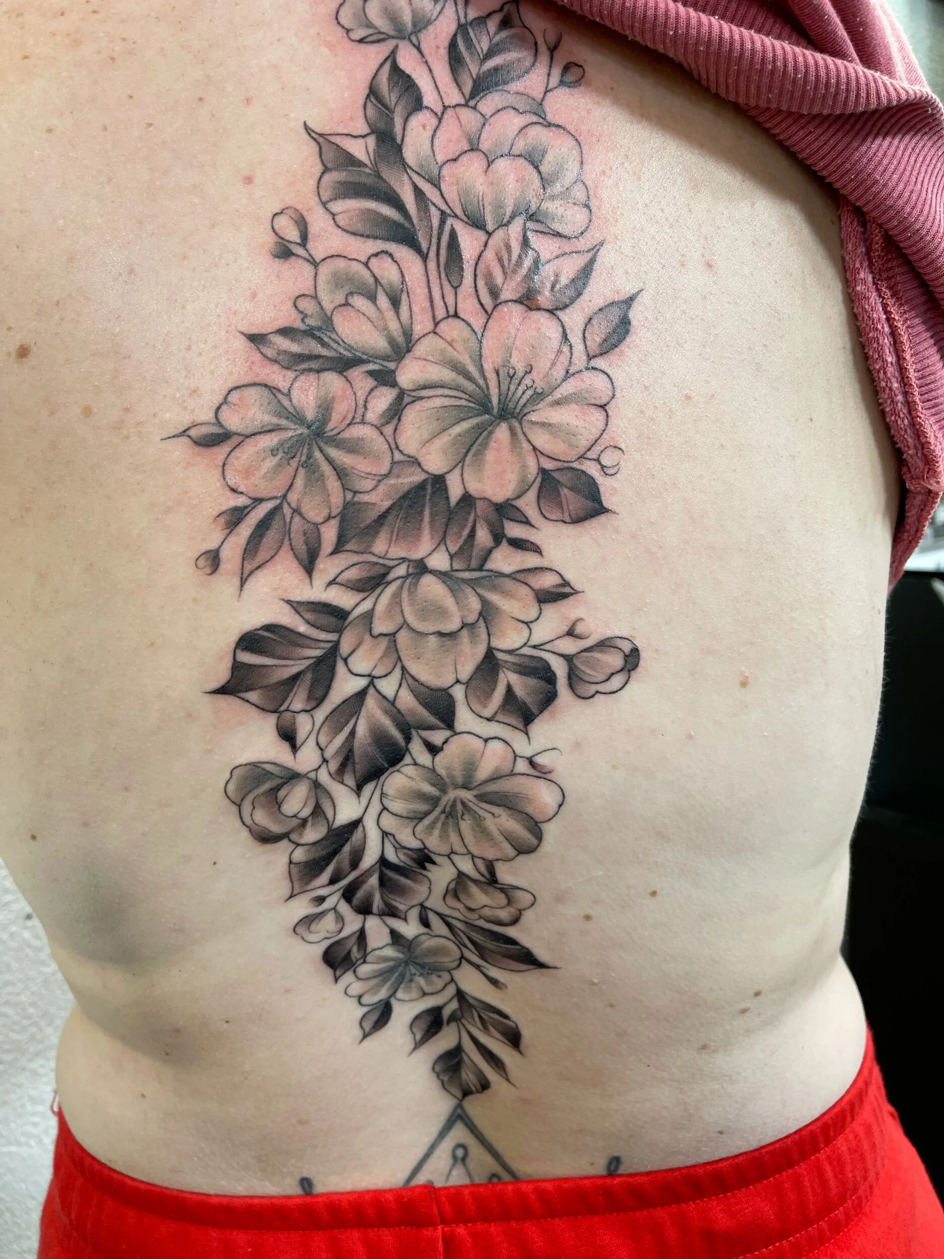 Black and grey floral tattoo on a person's side torso, featuring various flowers and leaves.