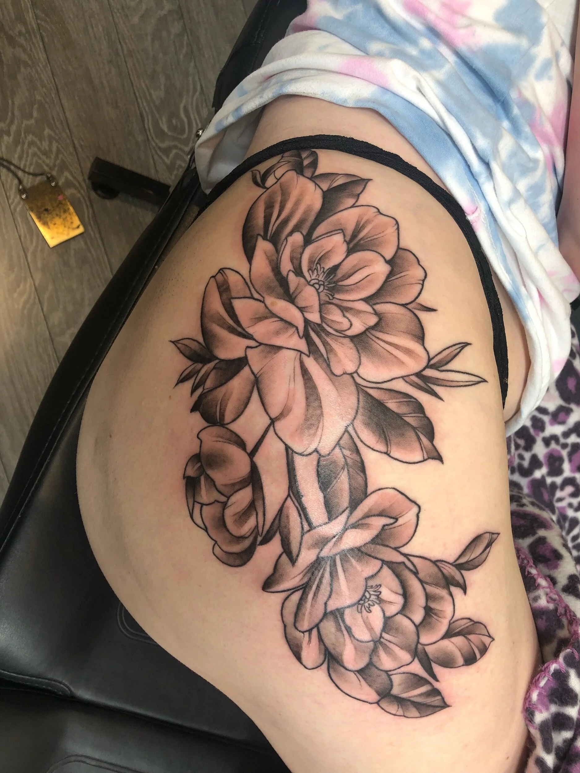 Large floral tattoo design on upper thigh, featuring black and gray shading of peonies and leaves.