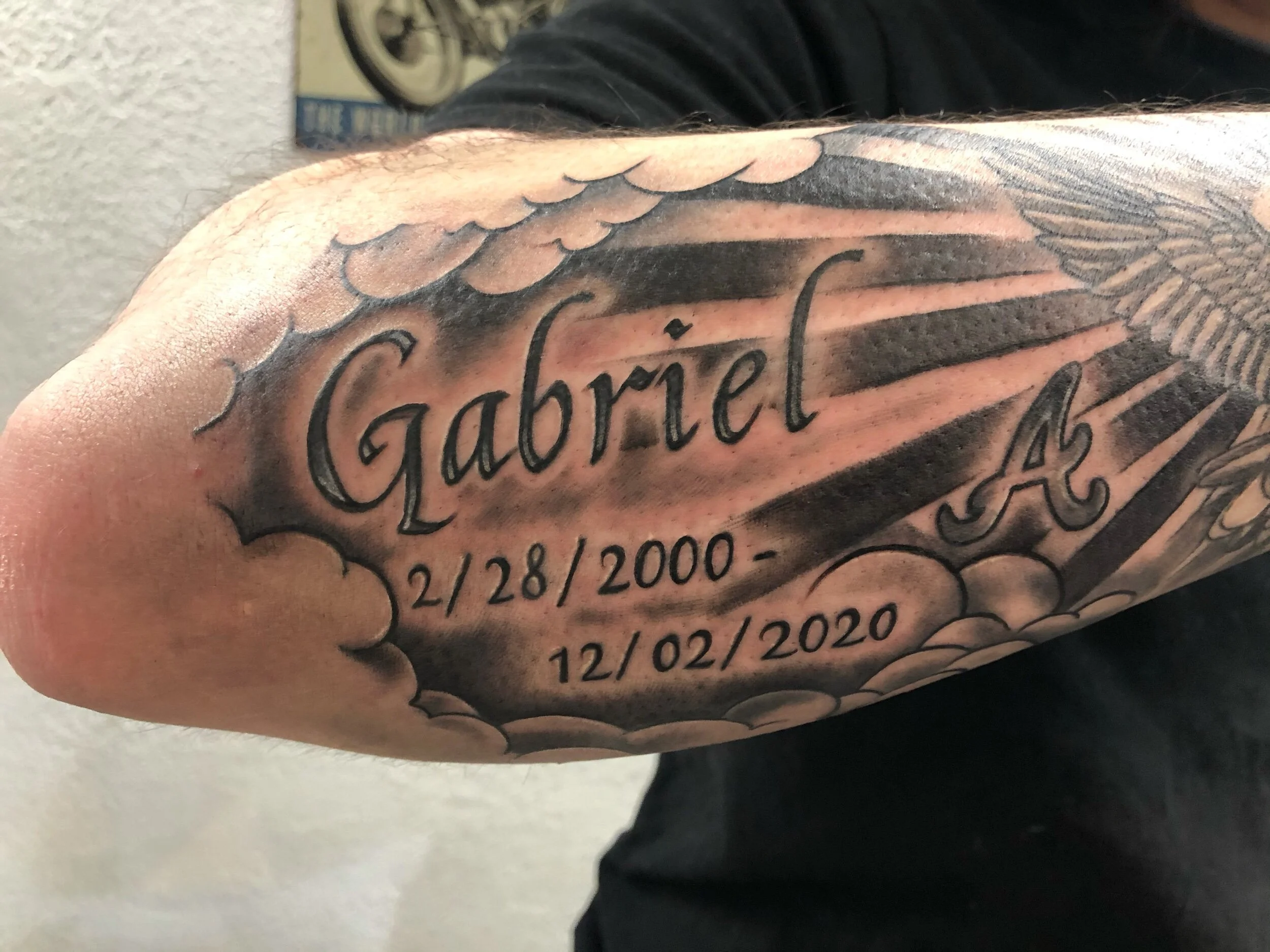 Black and gray tattoo on forearm with the name 'Gabriel,' dates '2/28/2000' and '12/02/2020,' clouds, sun rays, and an angel wing.