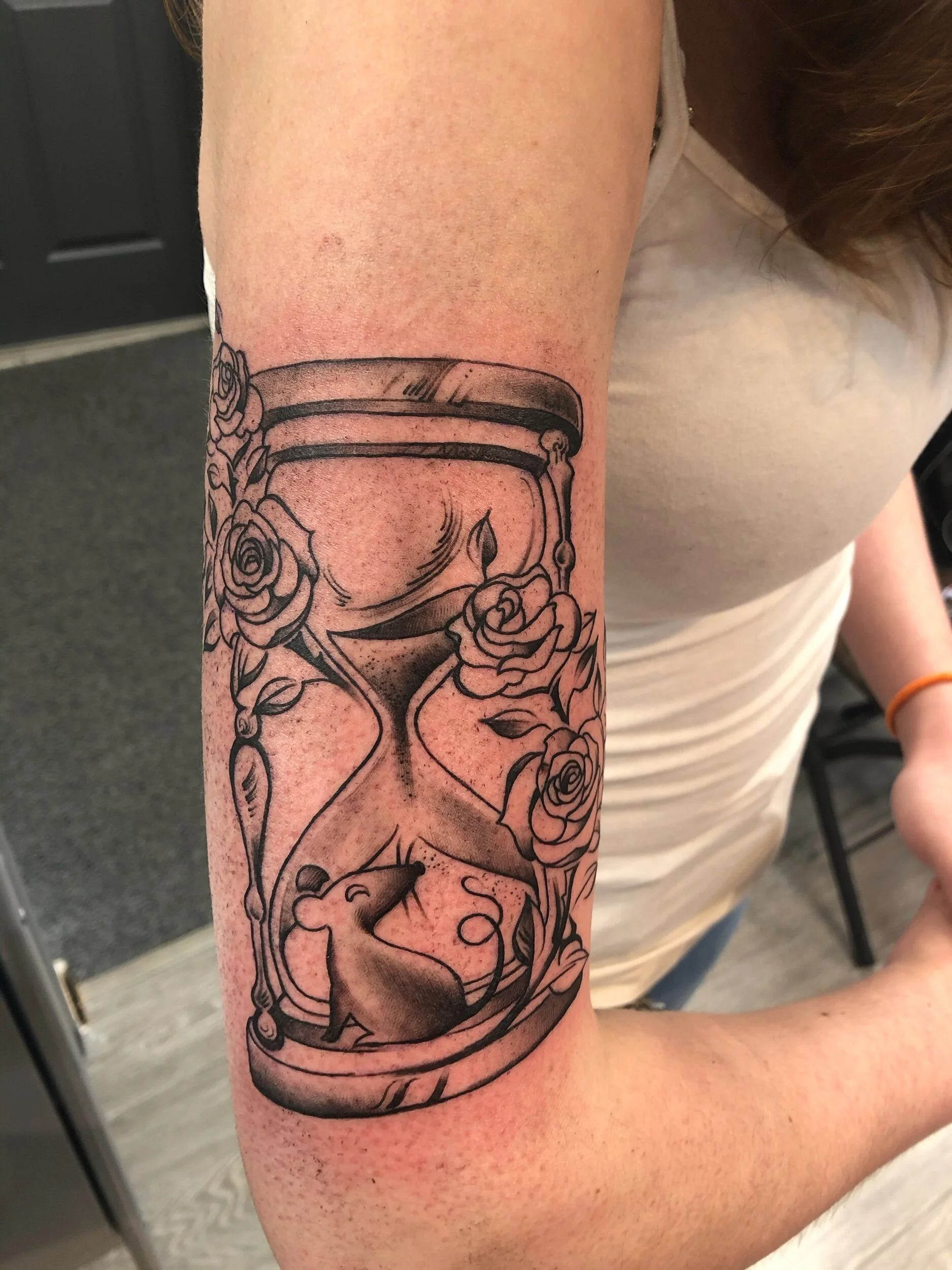 Tattoo featuring an hourglass with roses and a small mouse design on a person's arm.