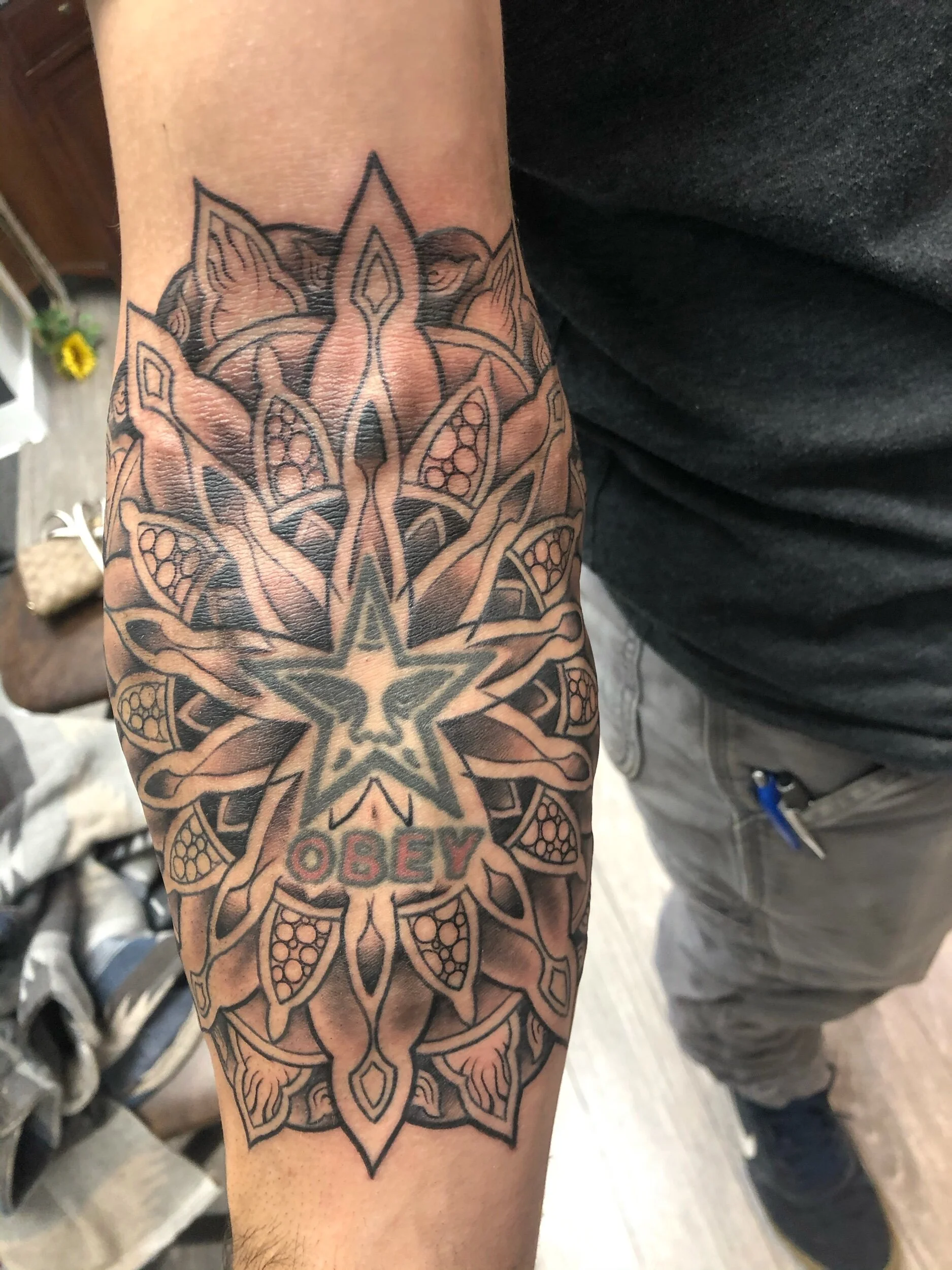 Forearm tattoo featuring intricate mandala design with a star and the word 'OBEY' in red letters.