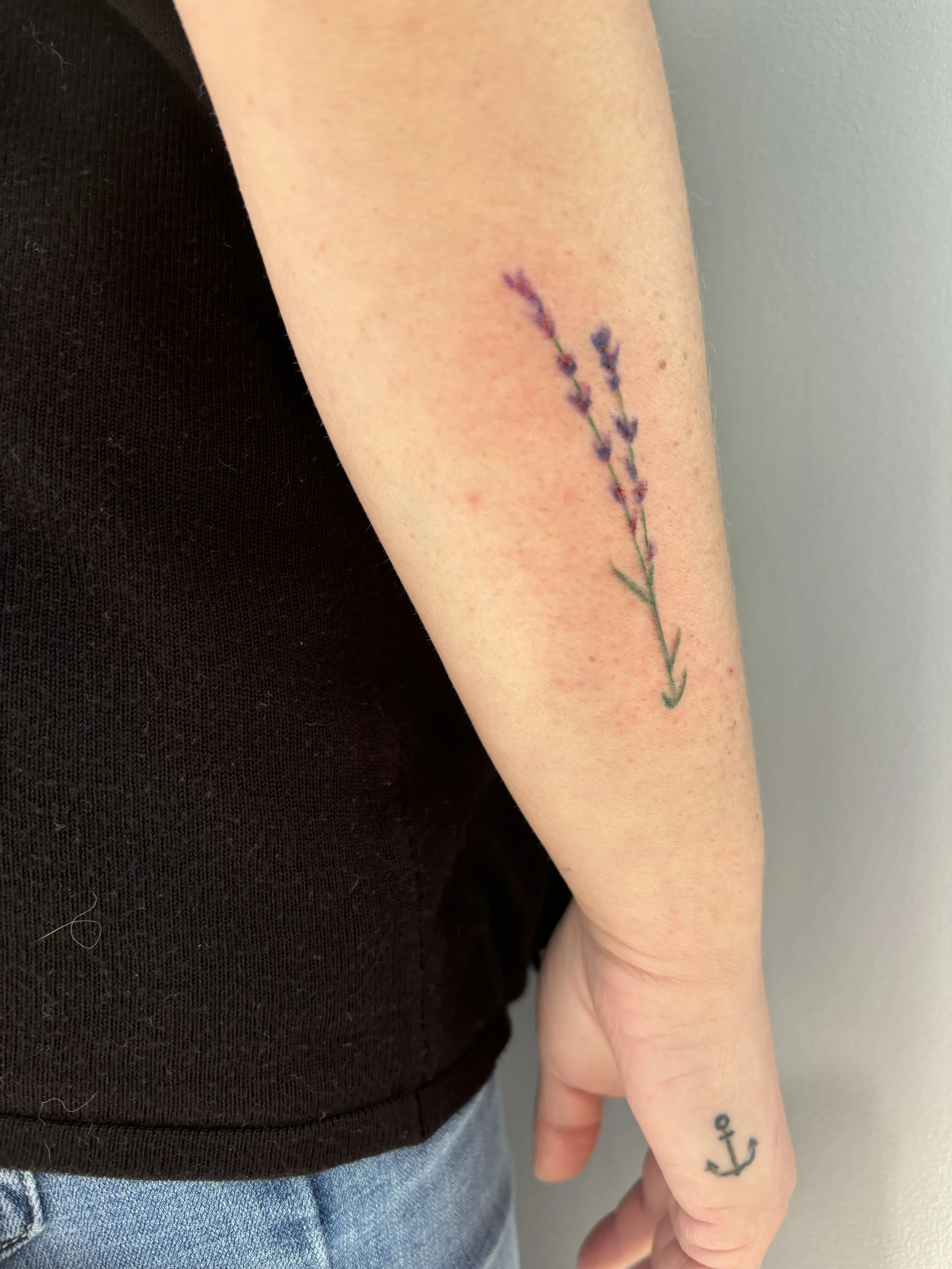 Tattoo of two lavender sprigs on the forearm, and a small black anchor on the finger.