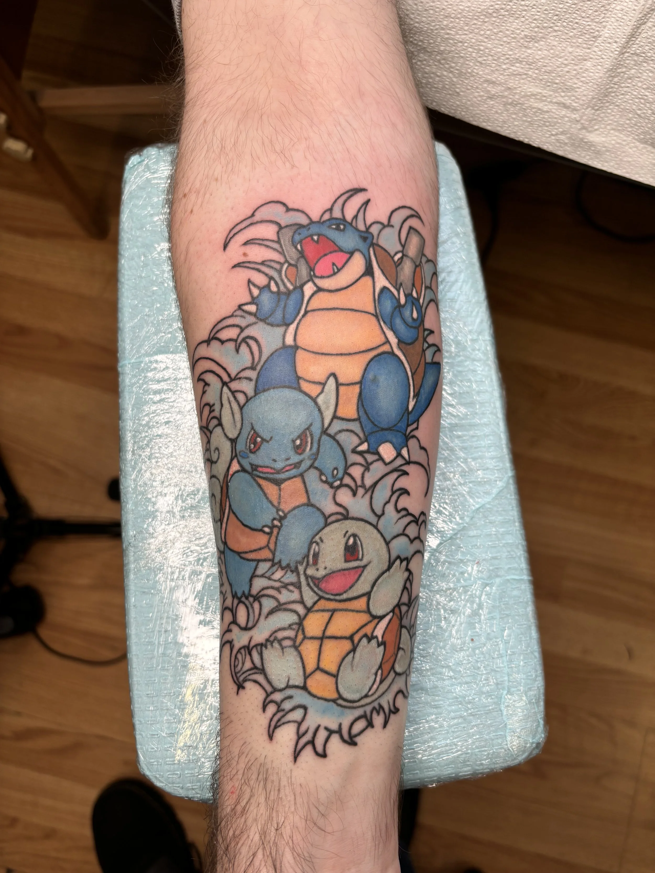 Colorful tattoo of Pokémon characters Blastoise, Venusaur, and Squirtle on a person's forearm with a cloud and wave background.