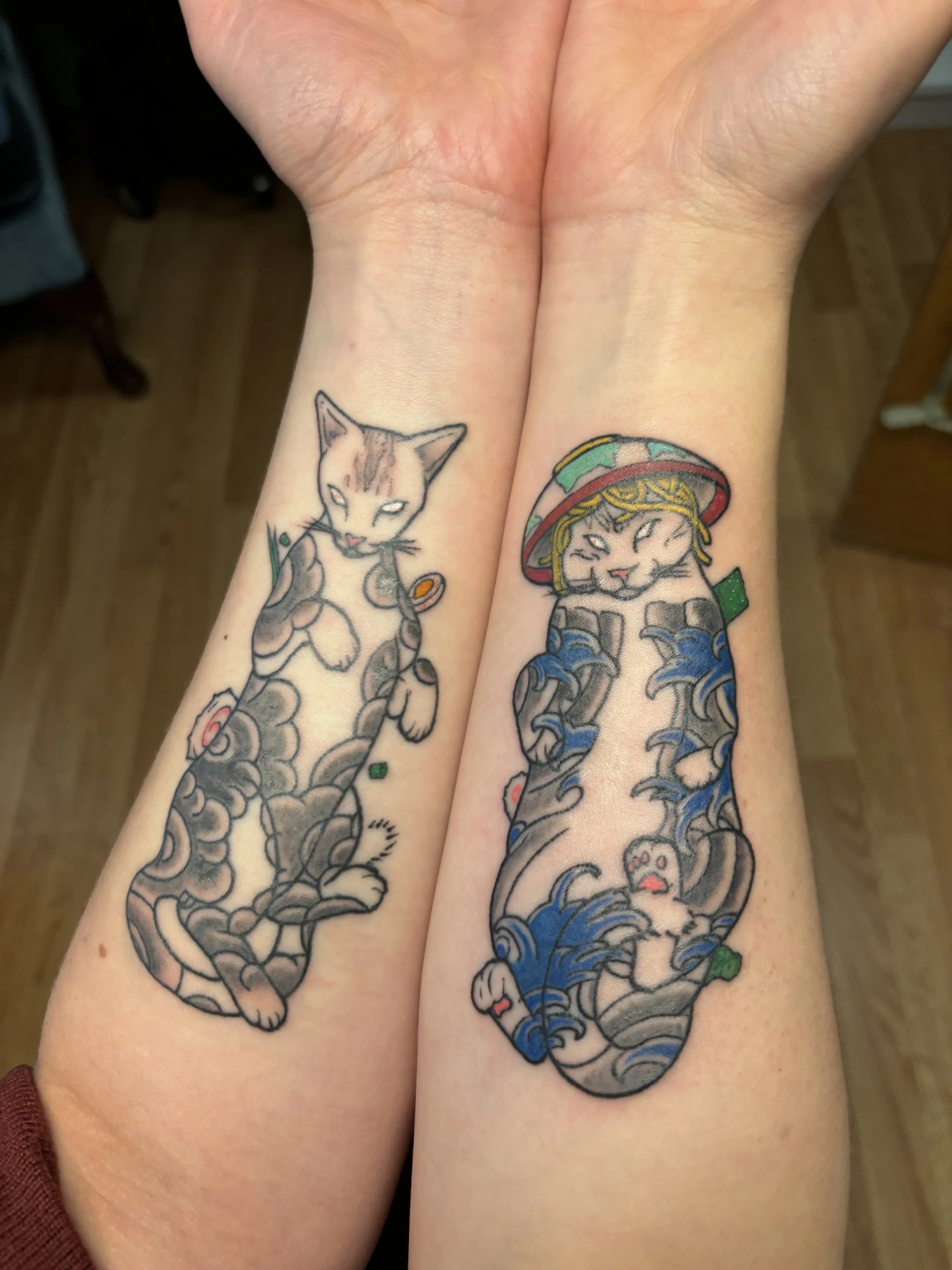 Two matching tattoos of stylized, cartoon-like cats with Japanese-inspired designs on the forearms.
