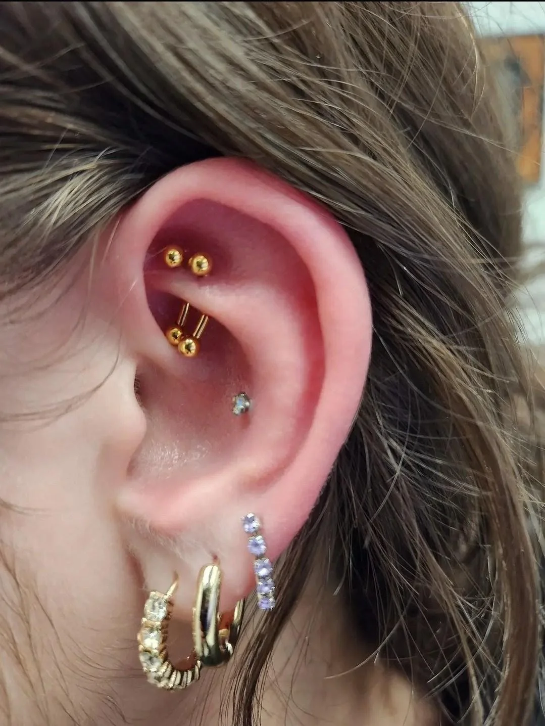 Close-up of a human ear with multiple piercings, including gold barbells, gemstone stud, and hoop earrings.