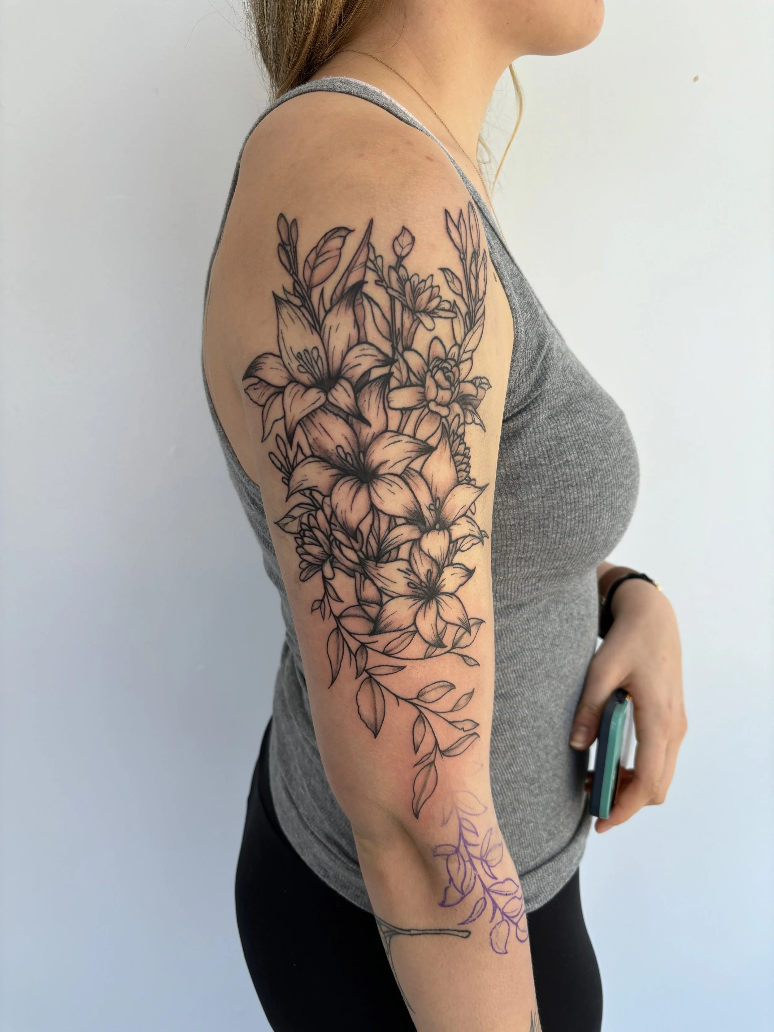 A woman with a detailed black outline floral tattoo on her upper arm, featuring lilies and various leaves, extending from her shoulder to her elbow.