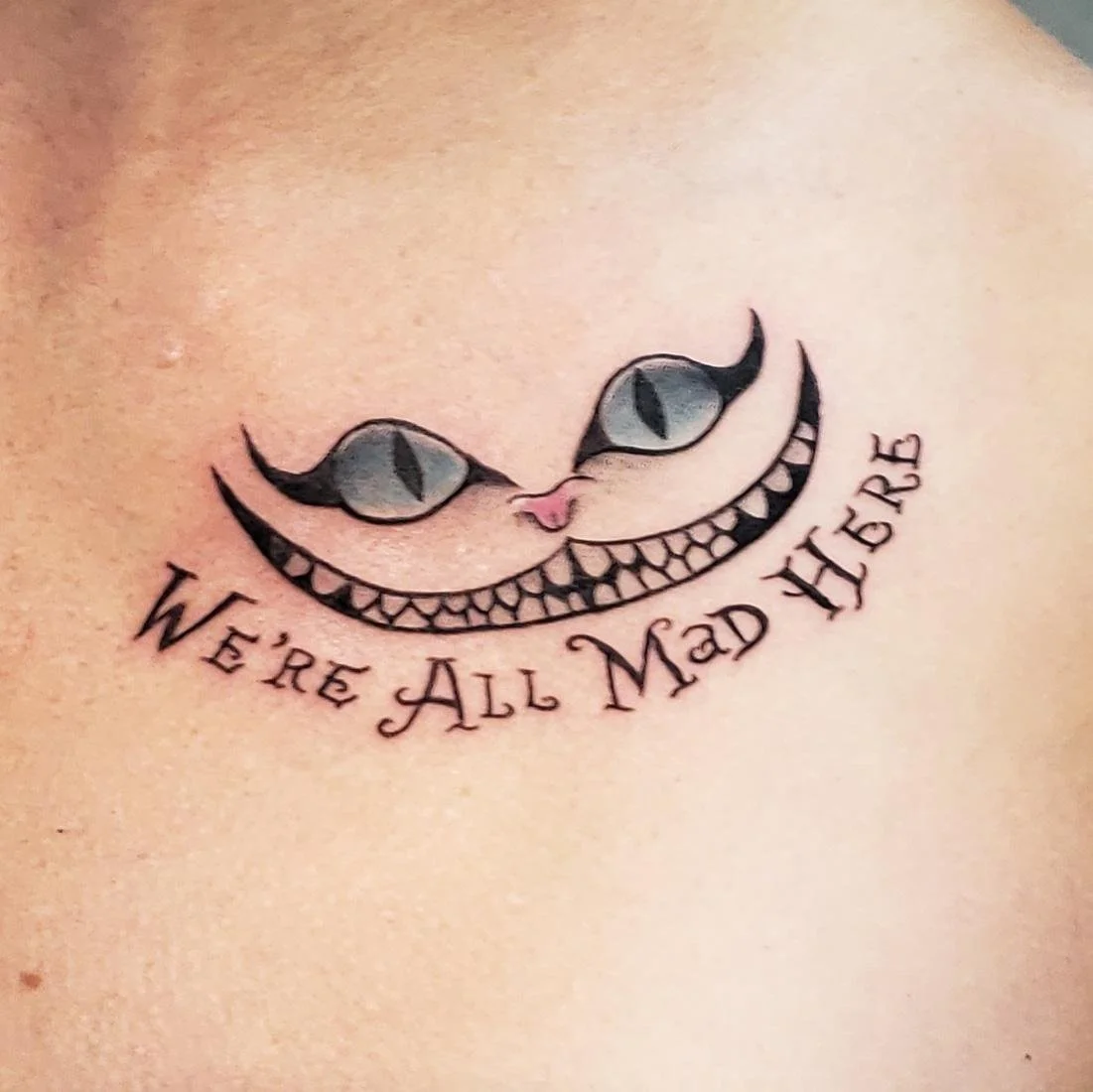 Tattoo of a grinning cat's face with the phrase "We're All Mad Here."