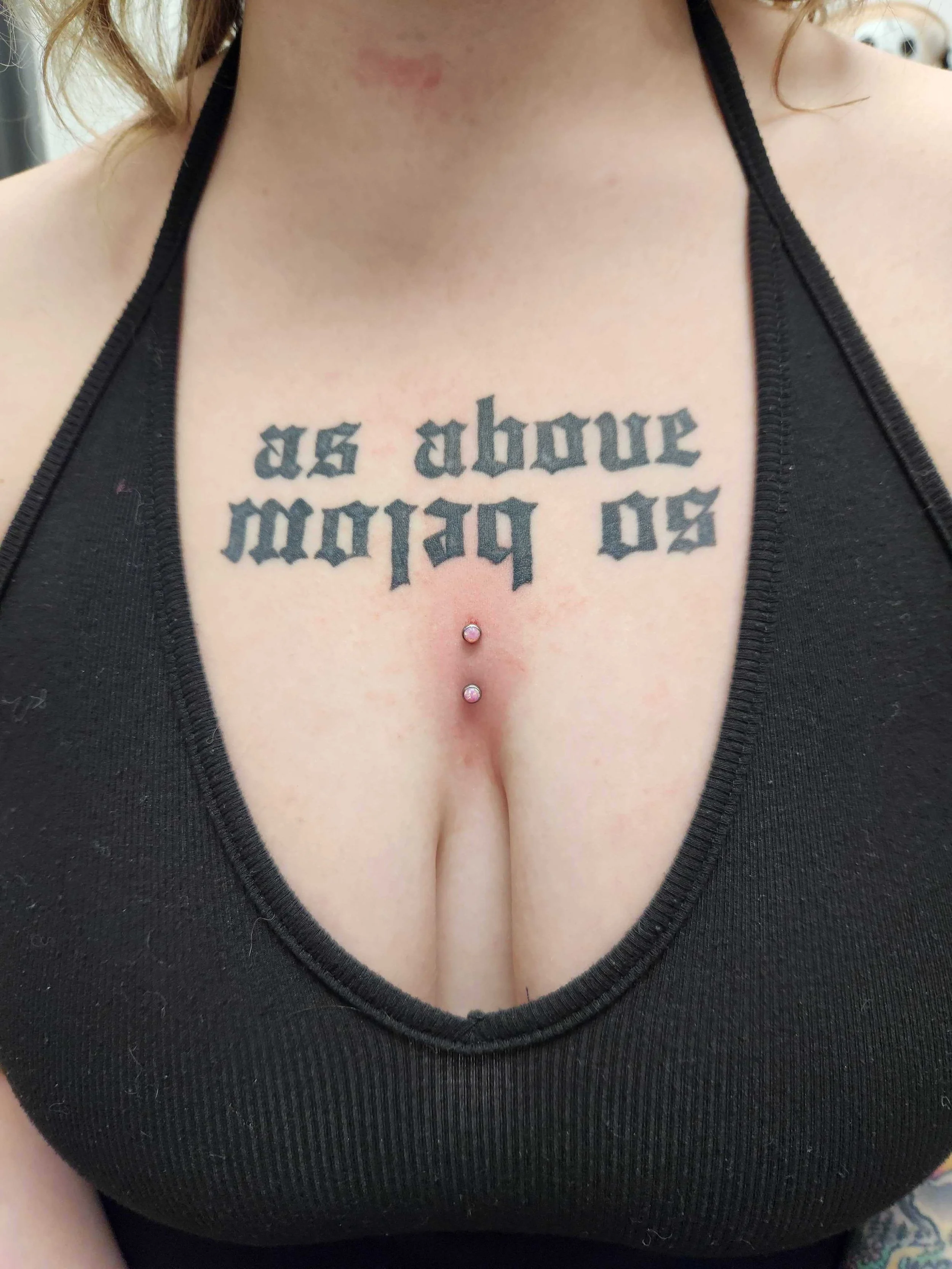A close-up of a woman's chest with a black halter top, featuring a tattoo in Gothic script that reads 'as above so below,' and a dermal piercing adorned with two pink gemstones.
